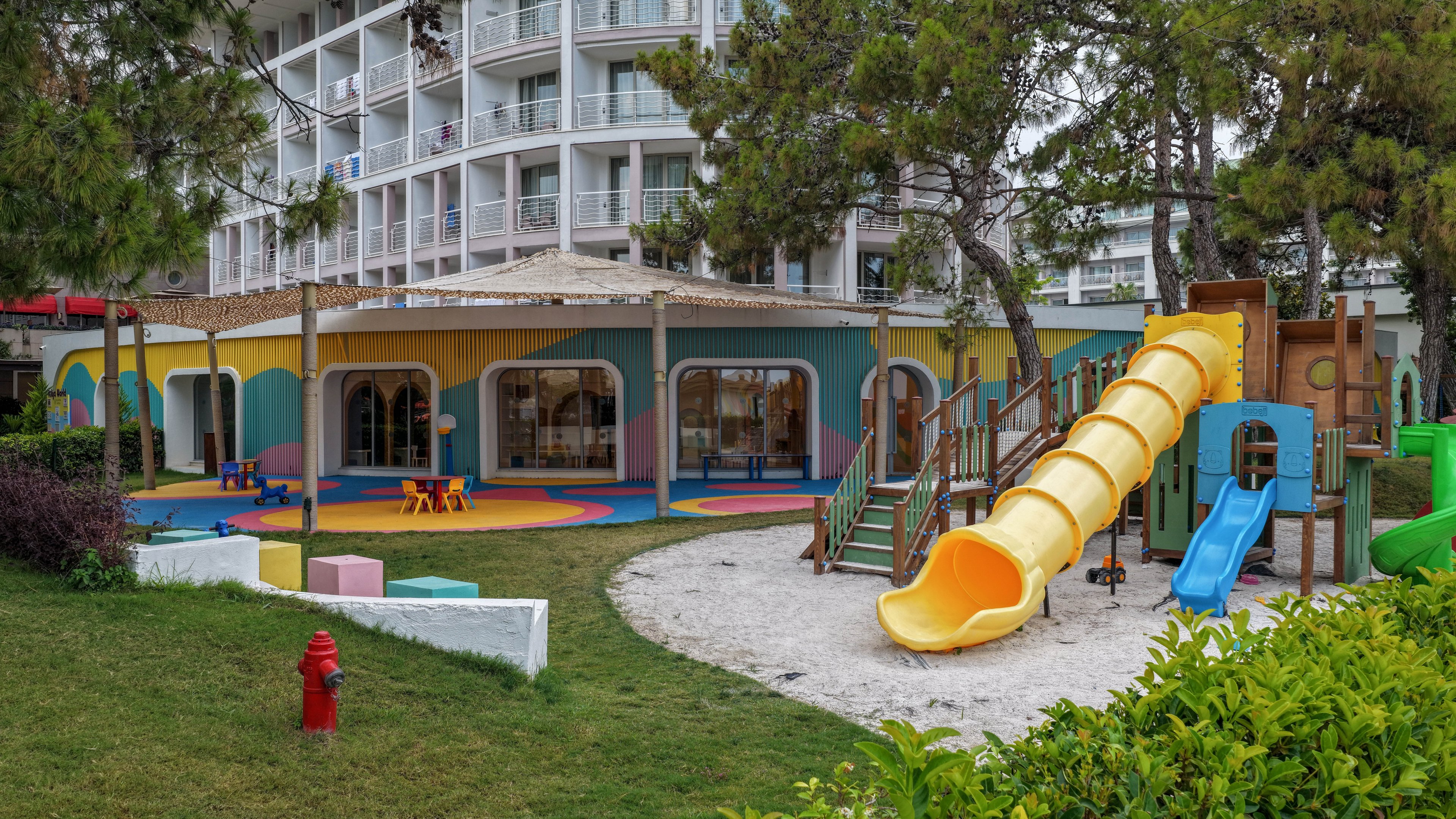 A vibrant outdoor playground is set in front of a modern multi-story hotel building. The play area features a large yellow tube slide, smaller slides, climbing structures, and colorful rubber flooring. Low trees and green grass surround the playground, creating a family-friendly atmosphere. A red fire hydrant stands in the foreground, adding a functional detail to the landscaped area.