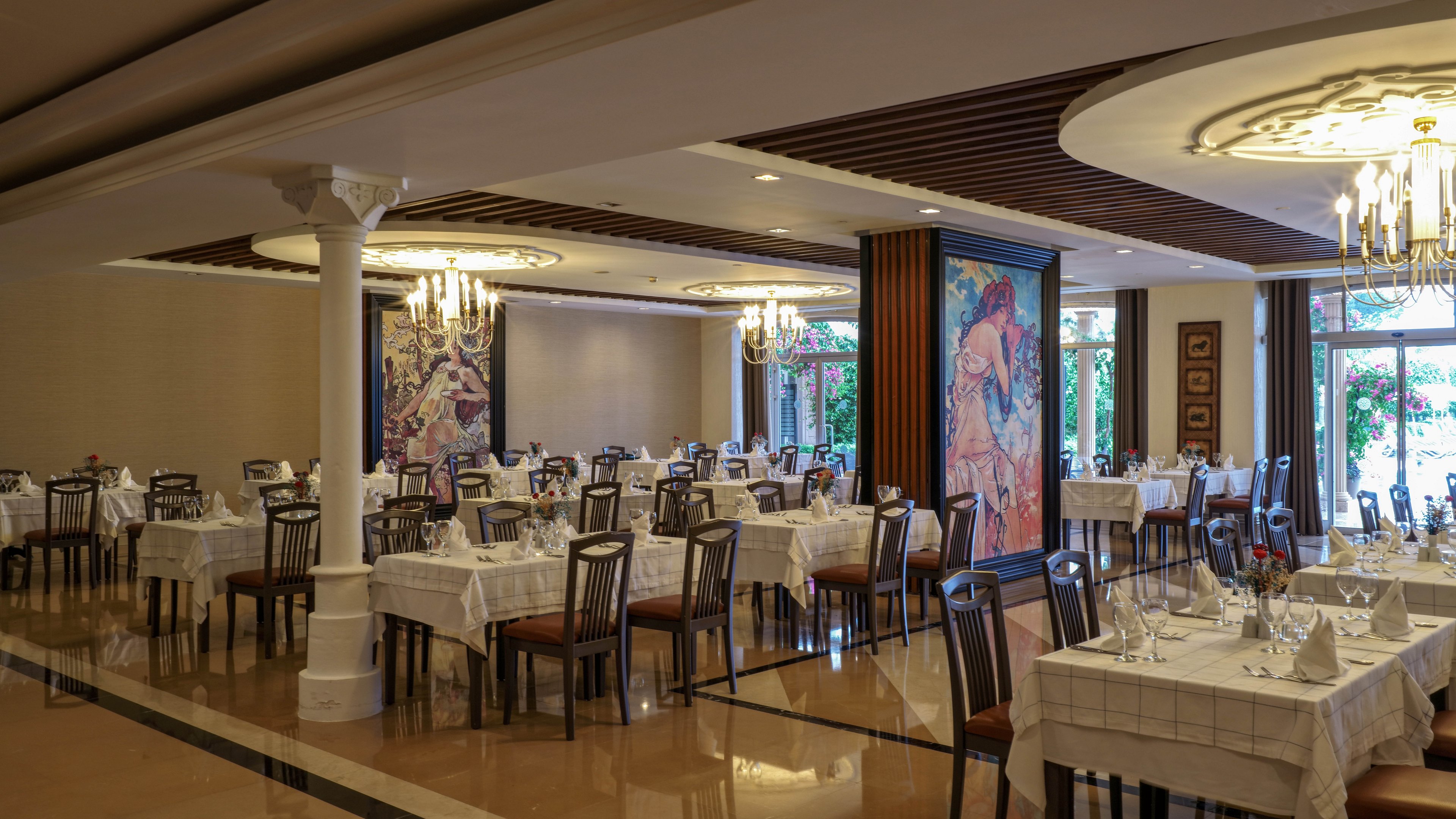 A spacious restaurant dining room is set with neatly arranged tables covered in white tablecloths. Wooden chairs surround each table, and wine glasses and cutlery are carefully placed, ready for guests. Chandeliers hang from the ceiling, casting warm light over the polished floor and decorative columns. Large colorful wall murals and windows with garden views add artistic and natural elements to the refined atmosphere.