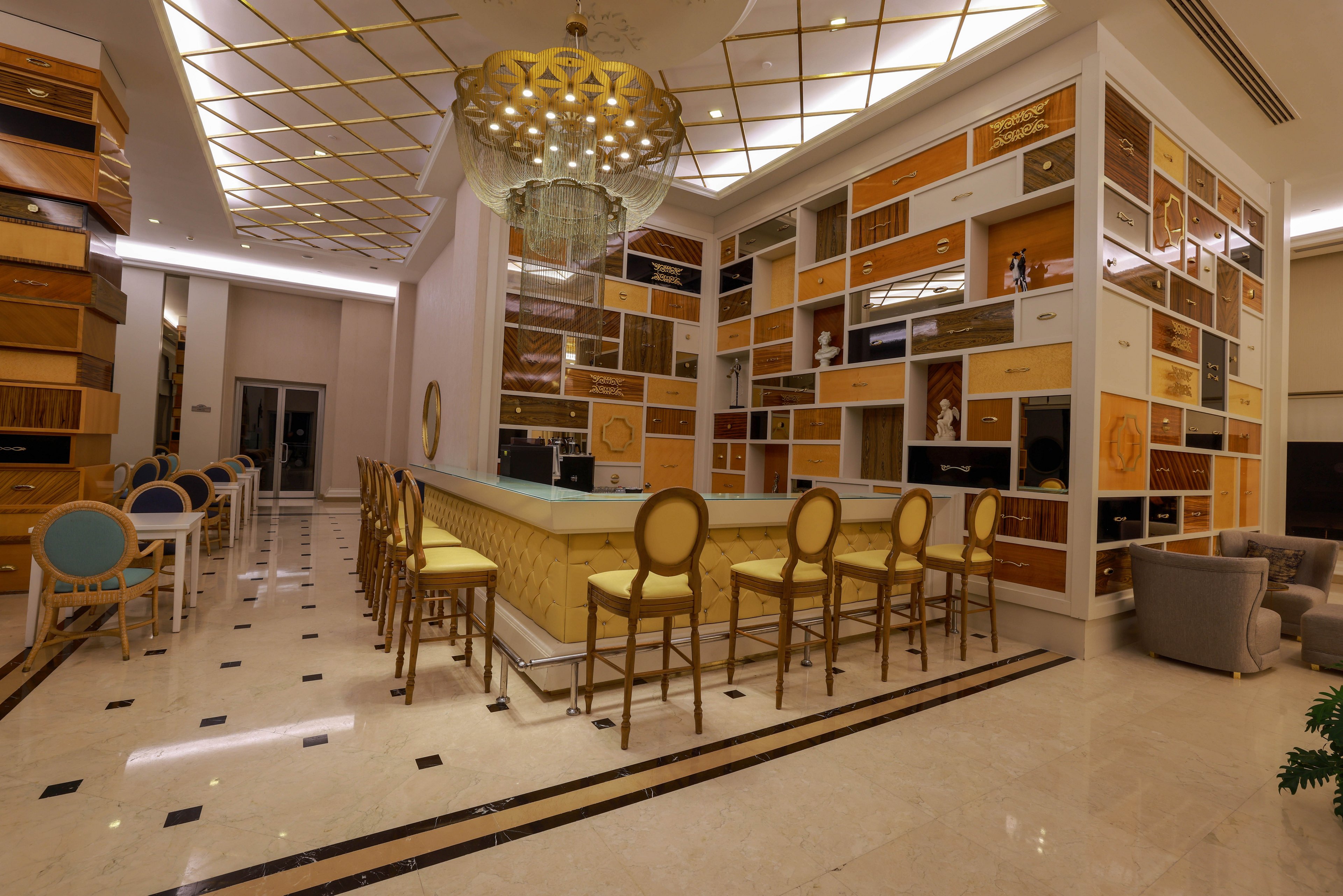 A stylish hotel bar features a long counter lined with high-backed stools in a warm golden tone. The surrounding walls are covered with geometric shelves and panels in white, beige, and wood finishes, creating a contemporary pattern. A large decorative chandelier hangs above the bar, casting soft light across the polished marble floor. In the background, additional seating with blue and white chairs and a lounge area with armchairs enhance the upscale, inviting atmosphere.