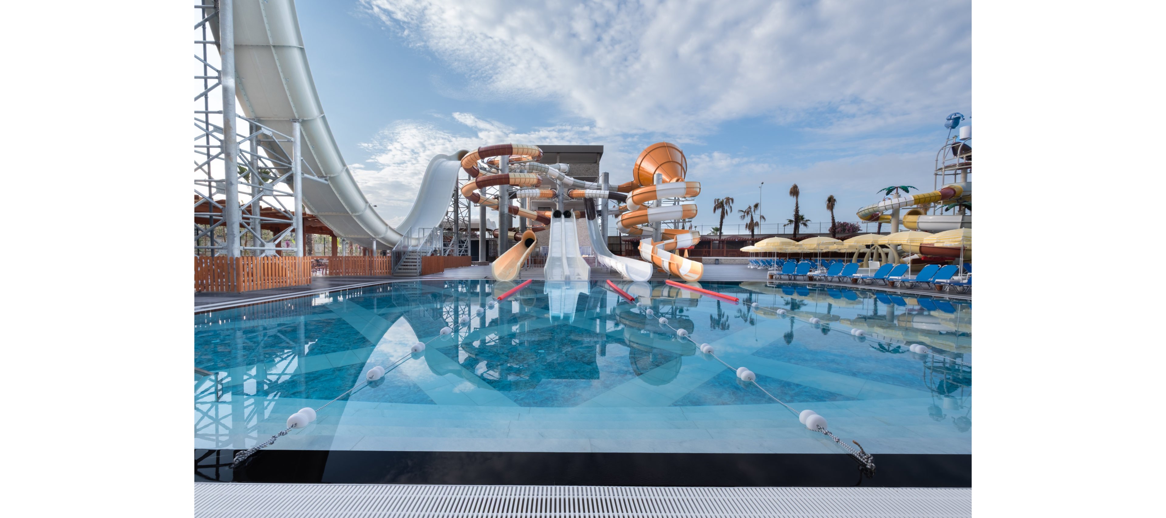 A large outdoor water park features multiple tall water slides leading into a clear blue pool. The scene is calm and empty, with no people visible in or around the water. Curved and straight slides in white and orange dominate the background structure. Palm trees, sun loungers, and umbrellas line the sides, under a partly cloudy sky.