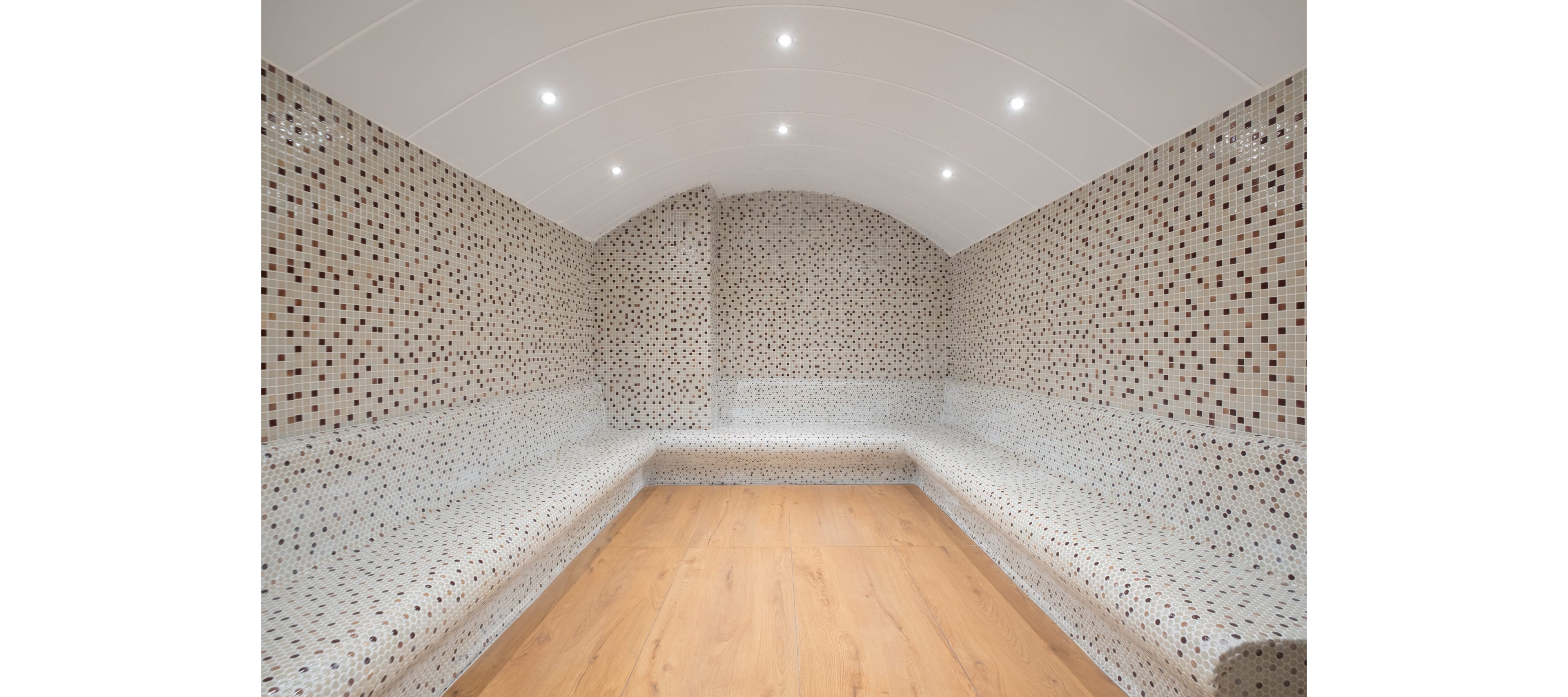 A contemporary steam room features curved walls and benches covered in small mosaic tiles. The ceiling is arched and fitted with recessed spotlights that evenly illuminate the space. Neutral tones of white, beige, and brown tiles contrast with the warm wooden floor. The clean, minimalist design suggests a spa or wellness environment focused on relaxation.