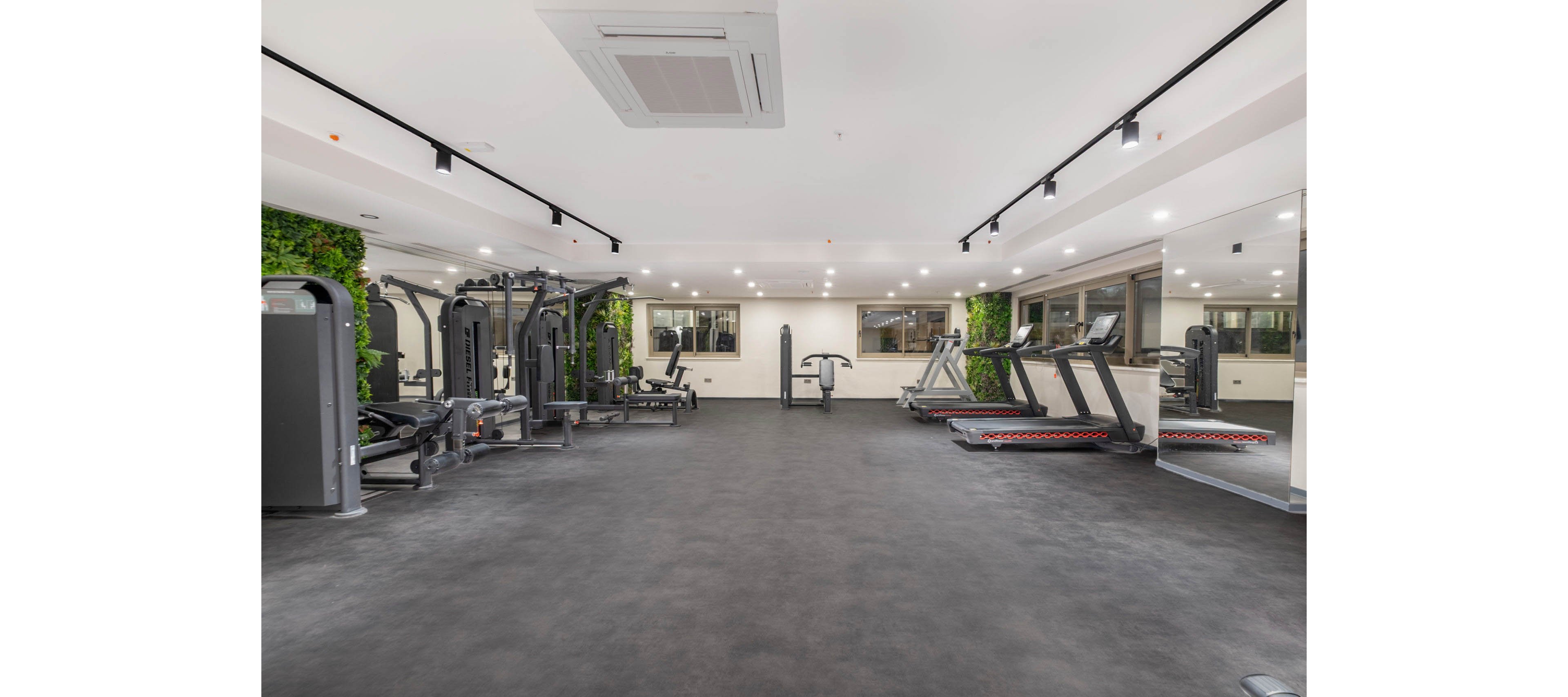 Spacious contemporary gym interior featuring various strength training machines and treadmills. The room is brightly lit with recessed ceiling lights and large windows along one wall. Green vertical plant panels add a natural accent to the otherwise minimalist white and gray space. The clean, uncluttered floor and absence of people emphasize readiness for workouts and fitness activities.