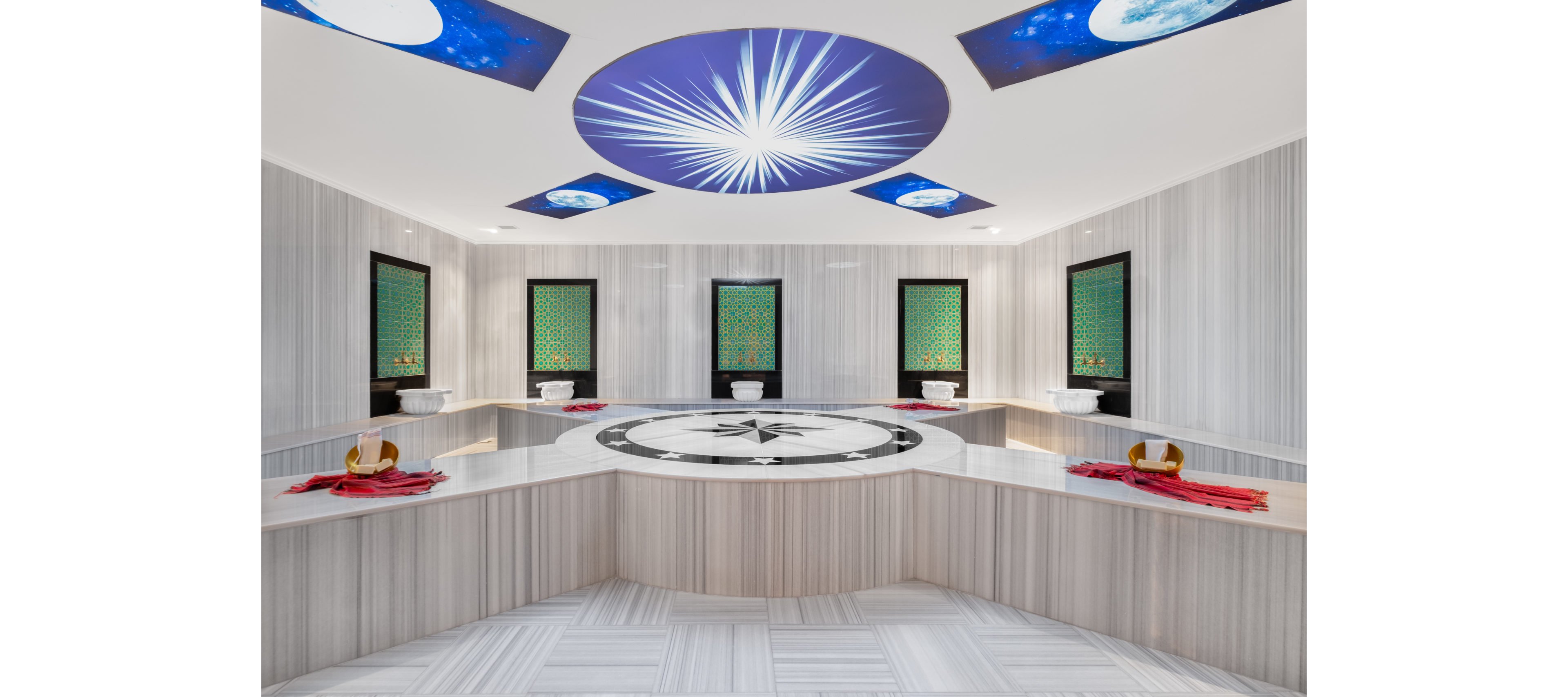 Spacious, contemporary hammam interior featuring a large central marble platform with a compass-like geometric design. The room is lined with light striped stone surfaces and individual washing stations along the walls, each prepared with red towels and traditional bowls. Decorative windows with intricate green patterns add cultural detail, while circular ceiling panels show starburst and moon imagery for a dramatic visual effect. The overall atmosphere is clean, luxurious, and serene, emphasizing symmetry and modern spa design.