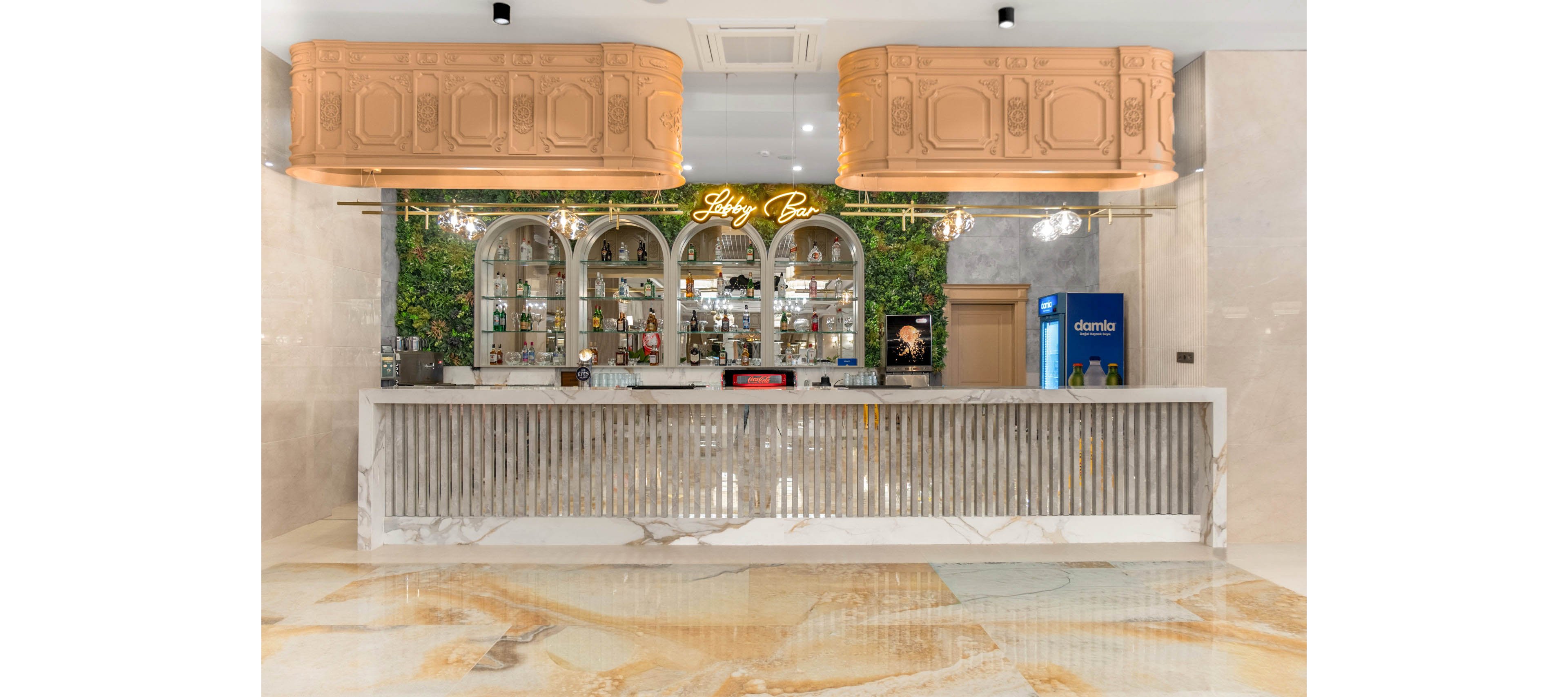 A stylish lobby bar features a long marble counter with vertical slats and a polished floor in warm beige tones. Behind the counter, arched shelves display various bottles and glassware framed by lush green foliage. A neon sign reading "Lobby Bar" glows above the shelves, adding a modern touch to the classic decor. On the right side, a blue refrigerator with the word "damla" is visible, along with a small service area and door.