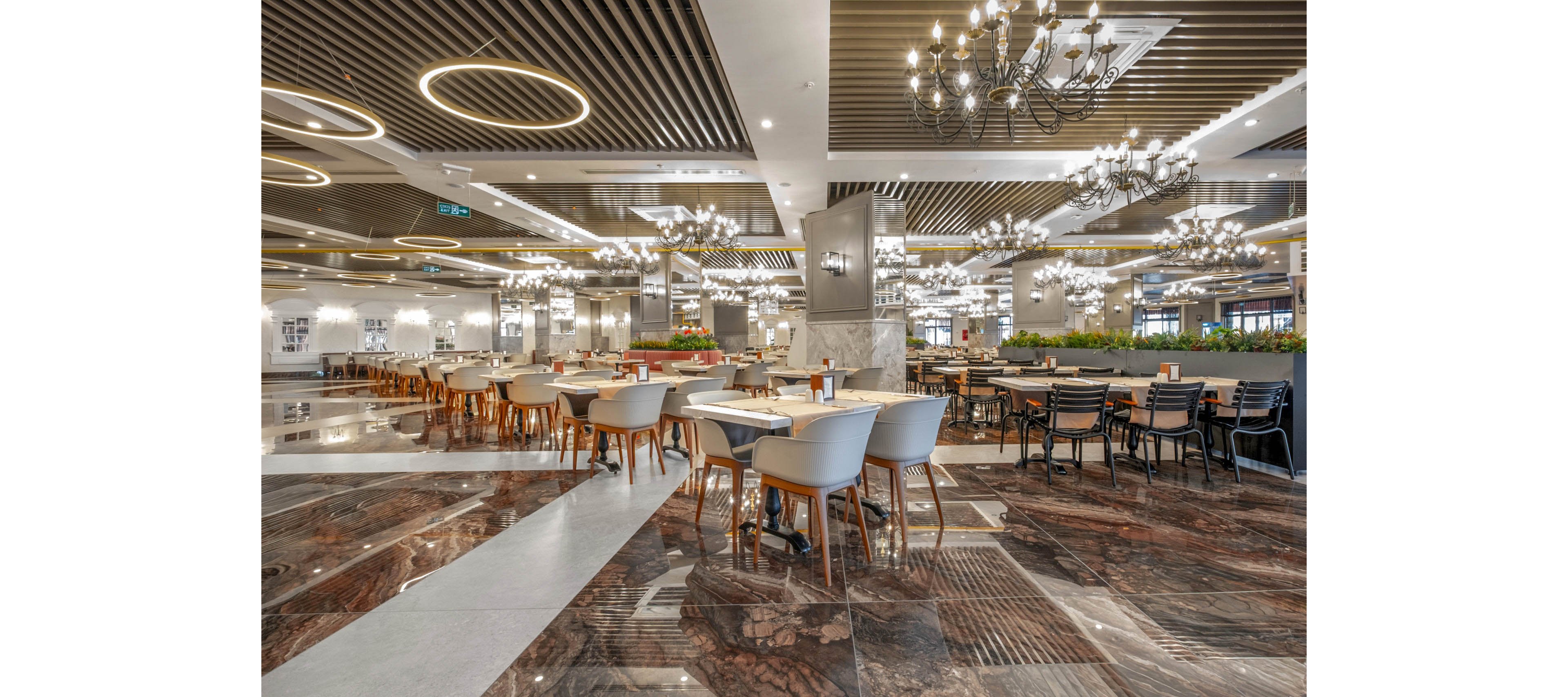 Spacious contemporary restaurant interior with rows of tables and chairs arranged neatly across a polished marble floor. Decorative chandeliers and circular ceiling lights create a bright, elegant atmosphere. Metal columns, wood slat ceilings, and planters with greenery add texture and warmth to the space. The dining area appears ready for guests, with table settings placed but no people present.