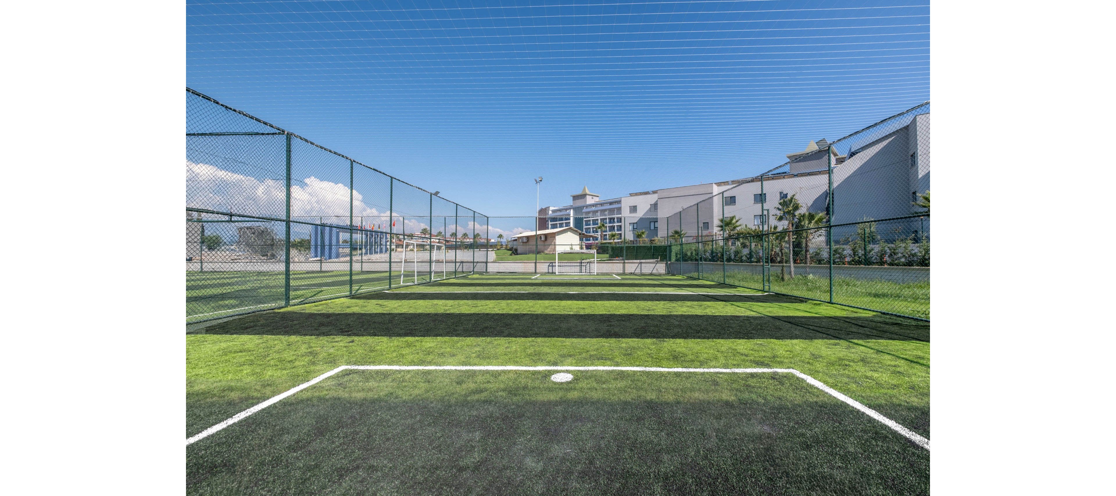 An empty outdoor soccer field is enclosed by tall chain-link fencing and netting under a clear blue sky. The artificial turf pitch features marked goal areas and a small white goal at the far end. Modern multi-story buildings and palm trees line the background, suggesting a recreational area within an urban or campus setting. Bright daylight and vivid green turf create a clean, inviting sports environment.