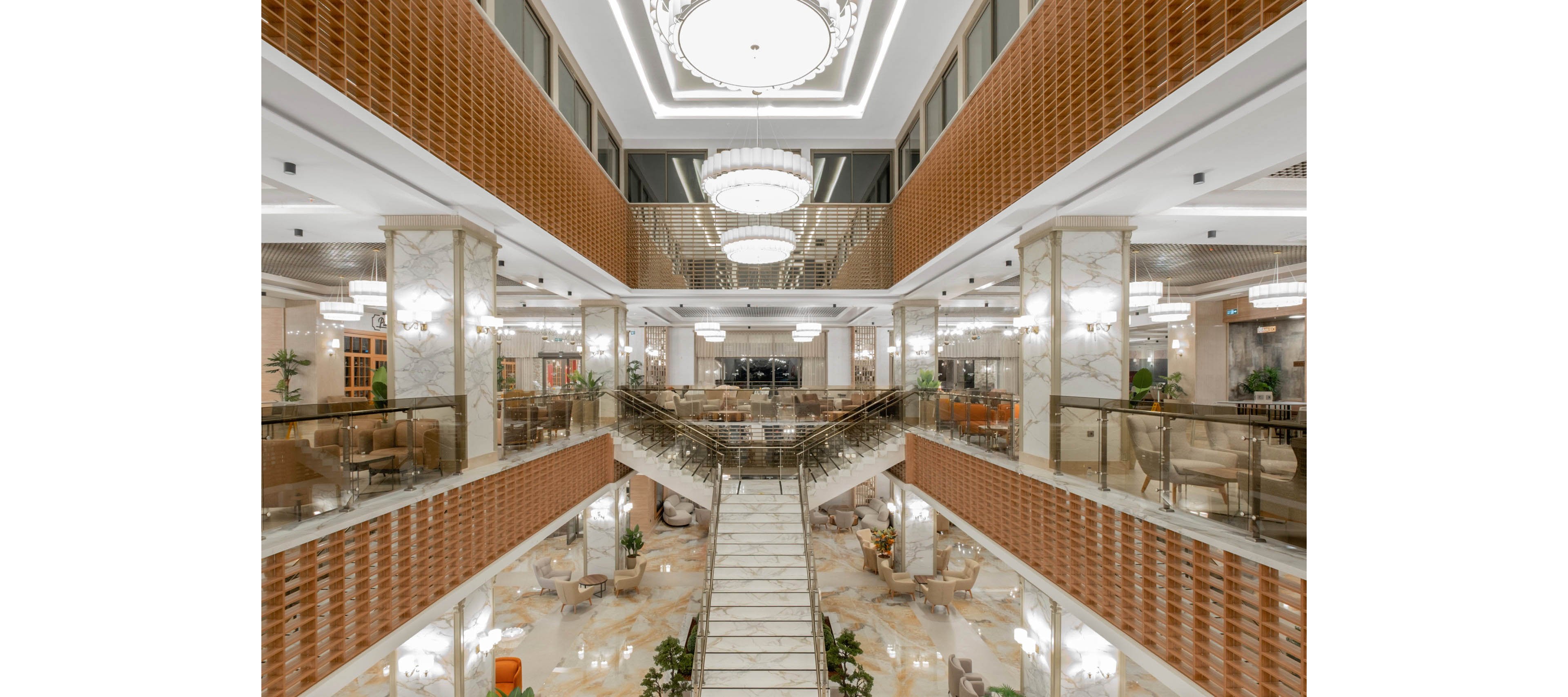 A spacious, multi-level hotel lobby features a grand central staircase leading down to an open seating area. Marble columns and floors, warm-toned wall panels, and glass railings create an elegant, contemporary interior. Large chandeliers and recessed ceiling lights illuminate the space, highlighting the symmetrical architectural design. Tables, chairs, and indoor plants are arranged neatly throughout the lobby, suggesting a comfortable social and waiting area.