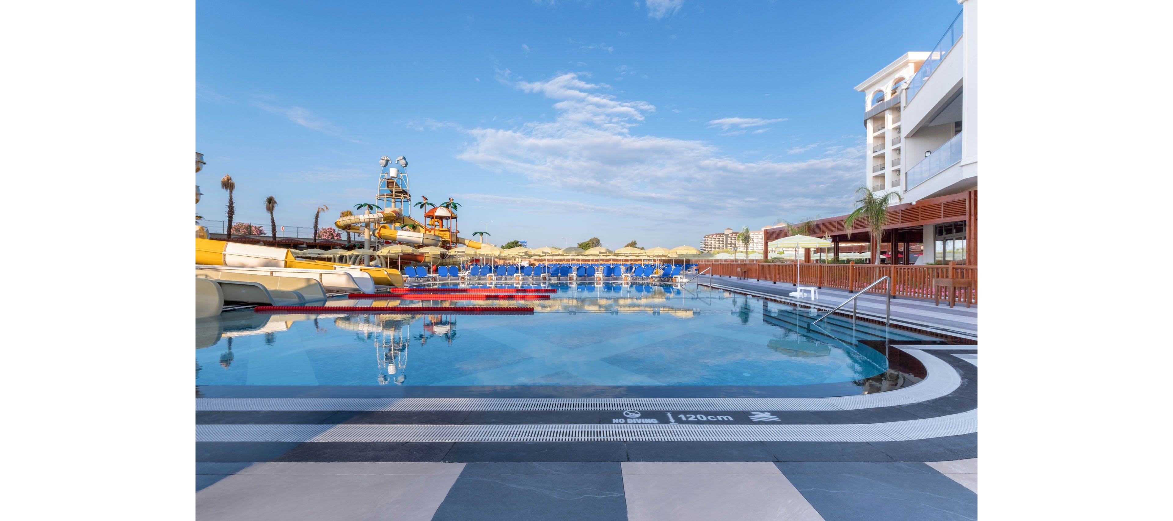 A large resort swimming pool reflects the clear blue sky and surrounding buildings. In the background, colorful water slides and numerous sun loungers with umbrellas create a lively vacation atmosphere. The poolside area features a metal handrail and tiled deck, with safety markings indicating a depth of 120 cm. Palm trees and multi-story hotel structures frame the scene, suggesting a modern holiday destination.