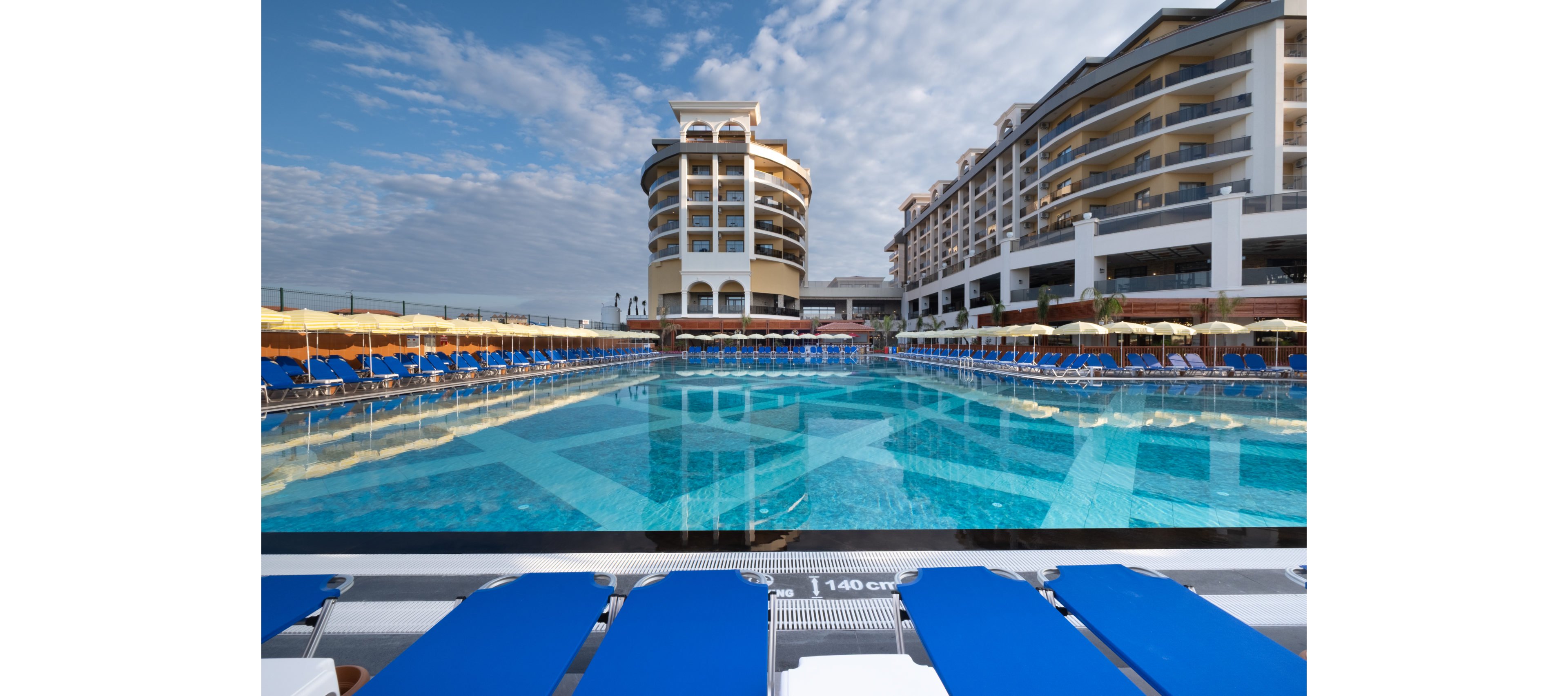 A large outdoor swimming pool dominates the foreground of a modern resort hotel complex. Rows of blue sun loungers and yellow umbrellas line the poolside, suggesting a relaxing vacation setting. The multi-story hotel buildings curve around the pool, reflecting in the clear blue water under a partly cloudy sky. A depth marker reading 1.40 cm is visible at the pool edge, adding detail to the scene.