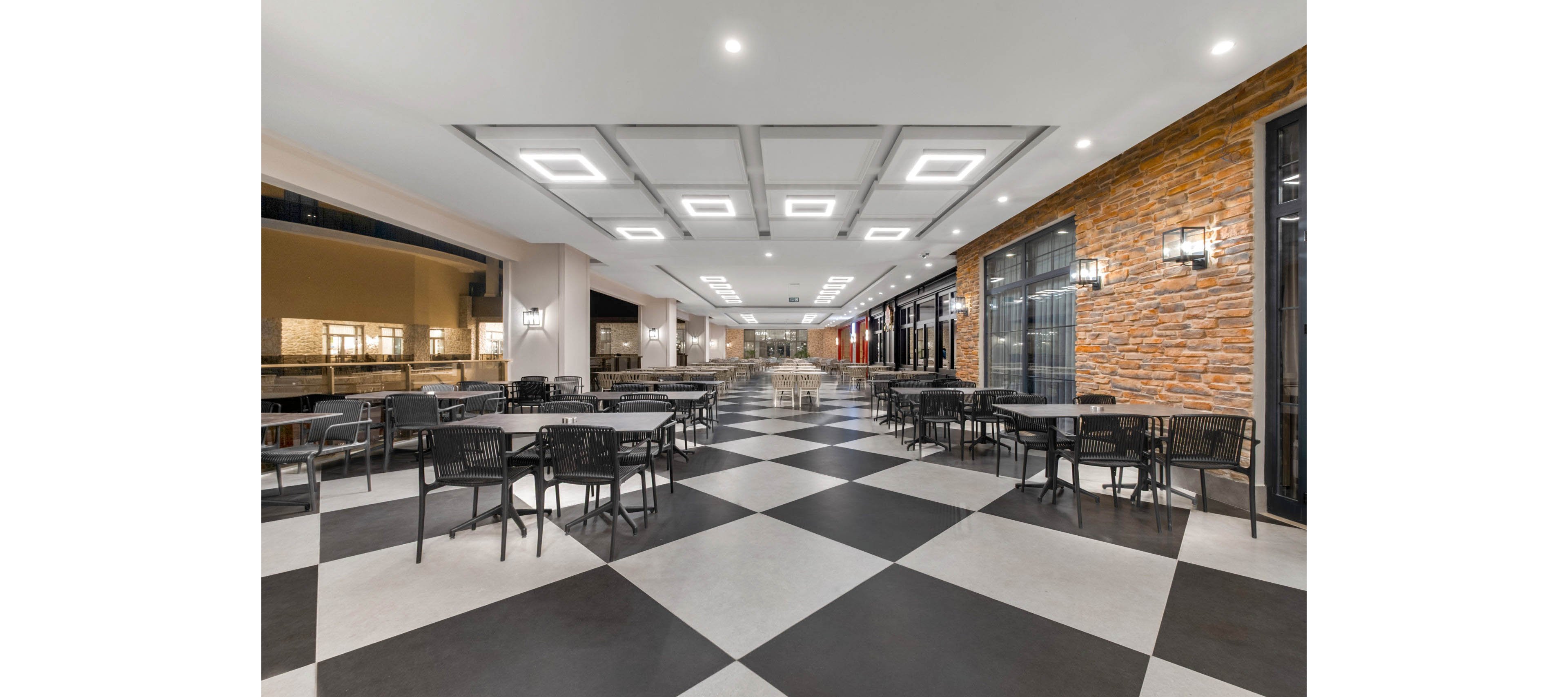 Spacious open-air dining area features rows of black tables and chairs arranged on a bold black-and-white checkered floor. The setting appears to be part of a contemporary restaurant or hotel, with large glass doors on one side and a textured stone wall. Bright recessed ceiling lights illuminate the space, creating a clean and inviting atmosphere. The area is empty of people, emphasizing the architectural design and layout of the seating.