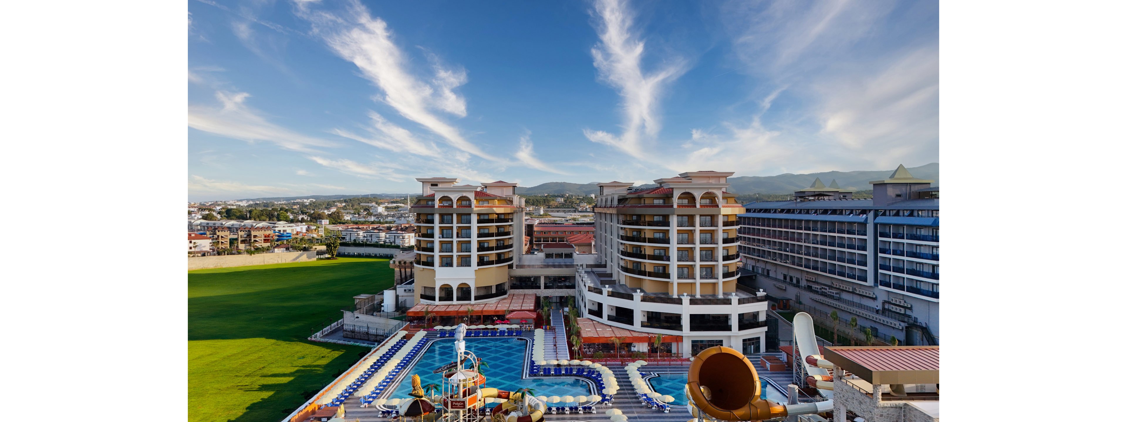 A large seaside resort complex features multiple modern hotel buildings surrounding expansive outdoor swimming pools. Numerous sun loungers line the pool decks, and colorful waterslides and play structures create a family-friendly water park area. In the background, a wide green field stretches toward distant hills under a bright blue sky with wispy clouds. The scene suggests a sunny vacation destination focused on leisure, relaxation, and outdoor fun.