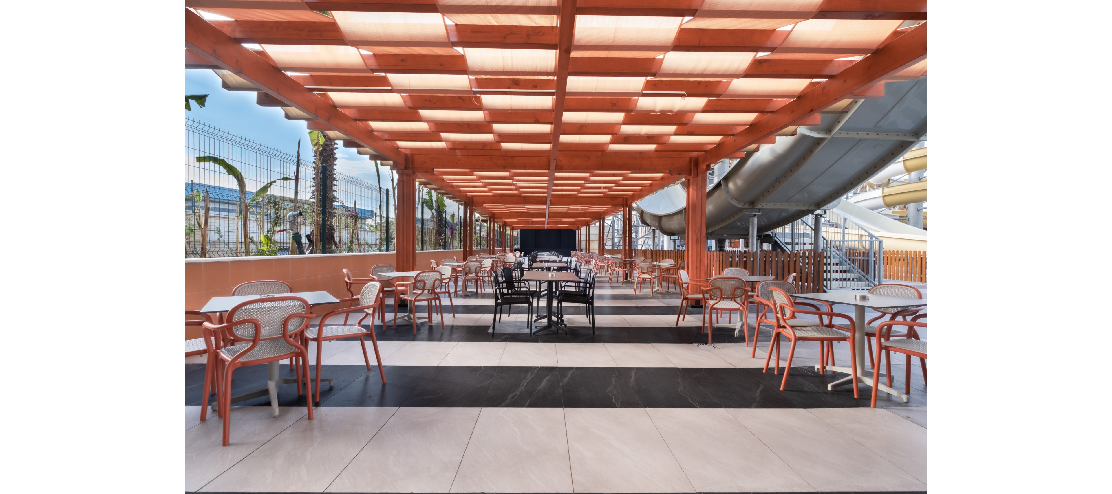 An open-air café terrace features rows of tables and chairs arranged beneath a modern wooden pergola. The space is bright and orderly, with tiled flooring in contrasting light and dark stripes leading toward the center. In the background, a water slide structure and fencing suggest the café is located within a leisure or resort area. The overall atmosphere is relaxed and inviting, suitable for casual dining or refreshments.
