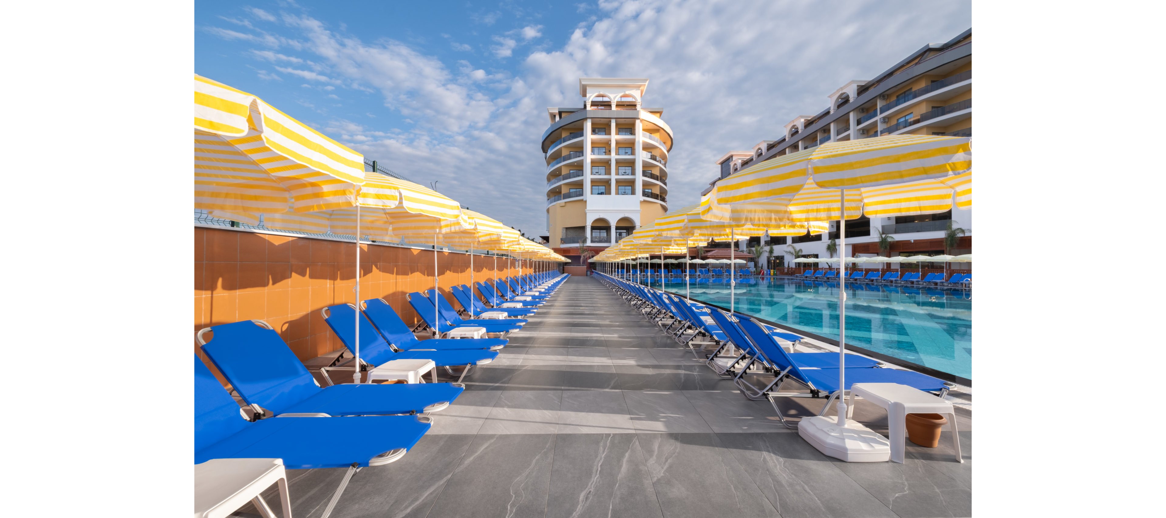 A row of bright blue sun loungers lines a resort poolside terrace beneath yellow and white striped umbrellas. The scene faces a round multi-story hotel building, with additional resort structures visible on the right. Clear blue water in the swimming pool reflects the sunny sky and scattered clouds. The area appears calm and empty, suggesting a relaxing vacation setting.