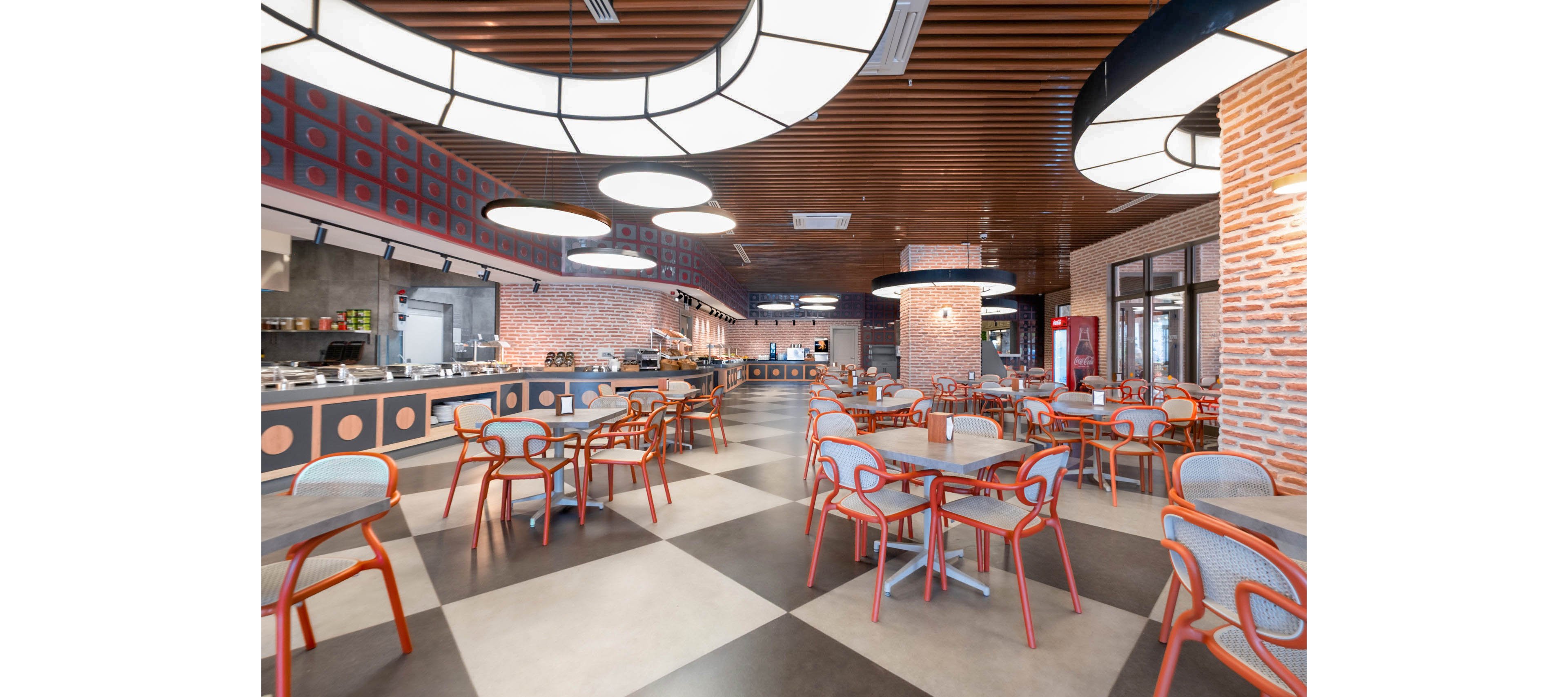 A spacious, contemporary cafeteria interior features rows of tables and chairs arranged on a geometric patterned floor. Warm brick walls and a wood-paneled ceiling create a cozy yet modern atmosphere. Large circular ceiling lights illuminate the open dining area and the self-service counter in the background. The space is empty of people, highlighting the clean, organized layout and architectural details.