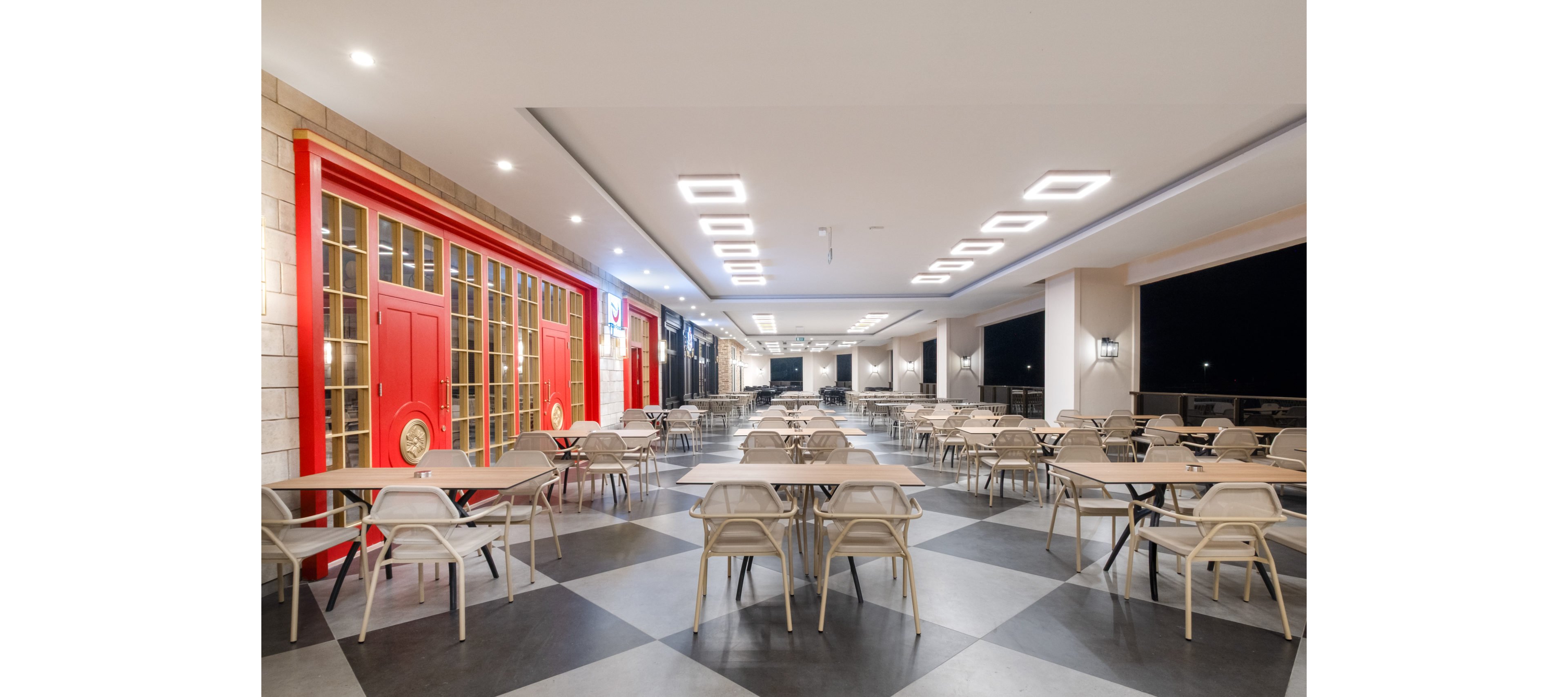 A spacious restaurant terrace features rows of empty tables and chairs arranged on a geometric checkered floor. Bright ceiling lights illuminate the area, creating a clean and modern atmosphere against the dark night sky. On the left side, large red doors and windows add a bold accent to the otherwise neutral color palette. The setting appears ready to welcome guests for evening dining or social gatherings.