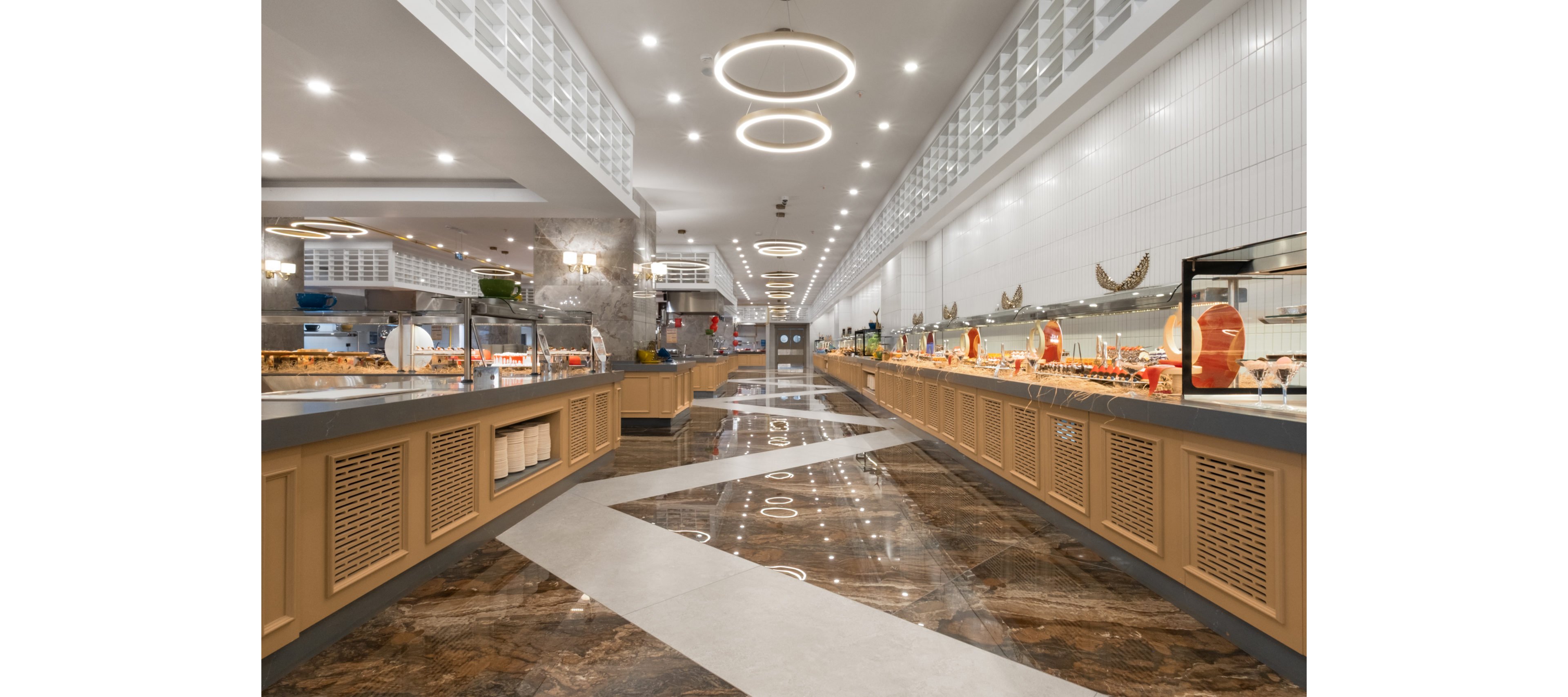 Wide-angle view of a contemporary buffet restaurant interior with long food counters on both sides. The setting features polished marble flooring with geometric patterns and sleek wooden cabinetry. Circular ceiling lights and bright recessed lighting create a clean, modern atmosphere. Various dishes and serving ware are arranged along the counters, suggesting a self-service dining experience.