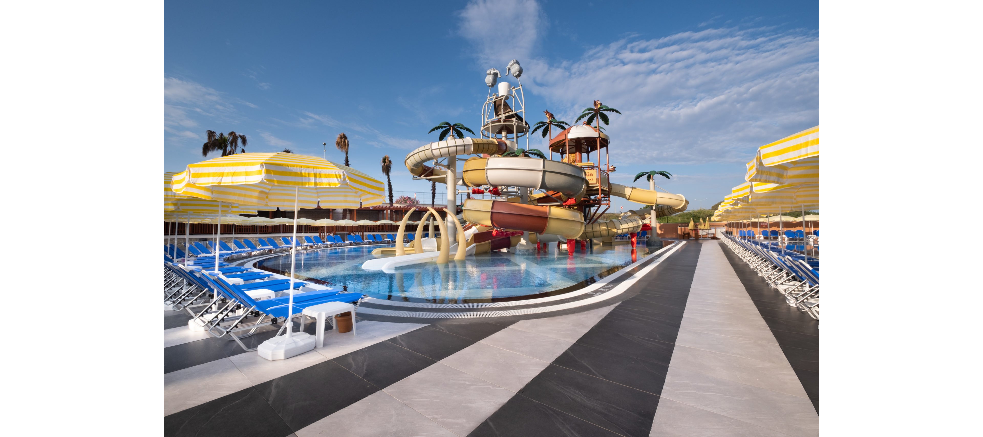 A vibrant outdoor water park features a central pool surrounded by large, twisting water slides and playful palm tree decorations. Numerous yellow-and-white striped umbrellas shade rows of blue lounge chairs lining both sides of the pool. The scene is bright and sunny, with clear blue sky and scattered clouds overhead. The overall atmosphere suggests a fun, family-friendly vacation destination.