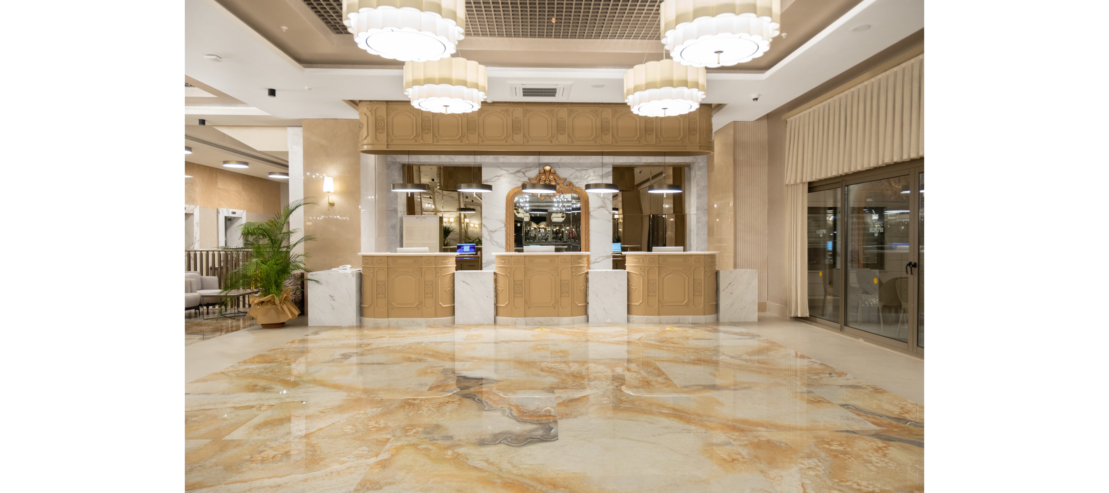 A spacious, elegant hotel lobby features a polished marble floor with warm beige and gold veining. The reception desk with multiple stations stands at the center, framed by decorative wood paneling and marble accents. Large pendant chandeliers hang from the ceiling, casting soft light across the room. Glass doors on the right and seating with plants on the left complete the upscale interior setting.