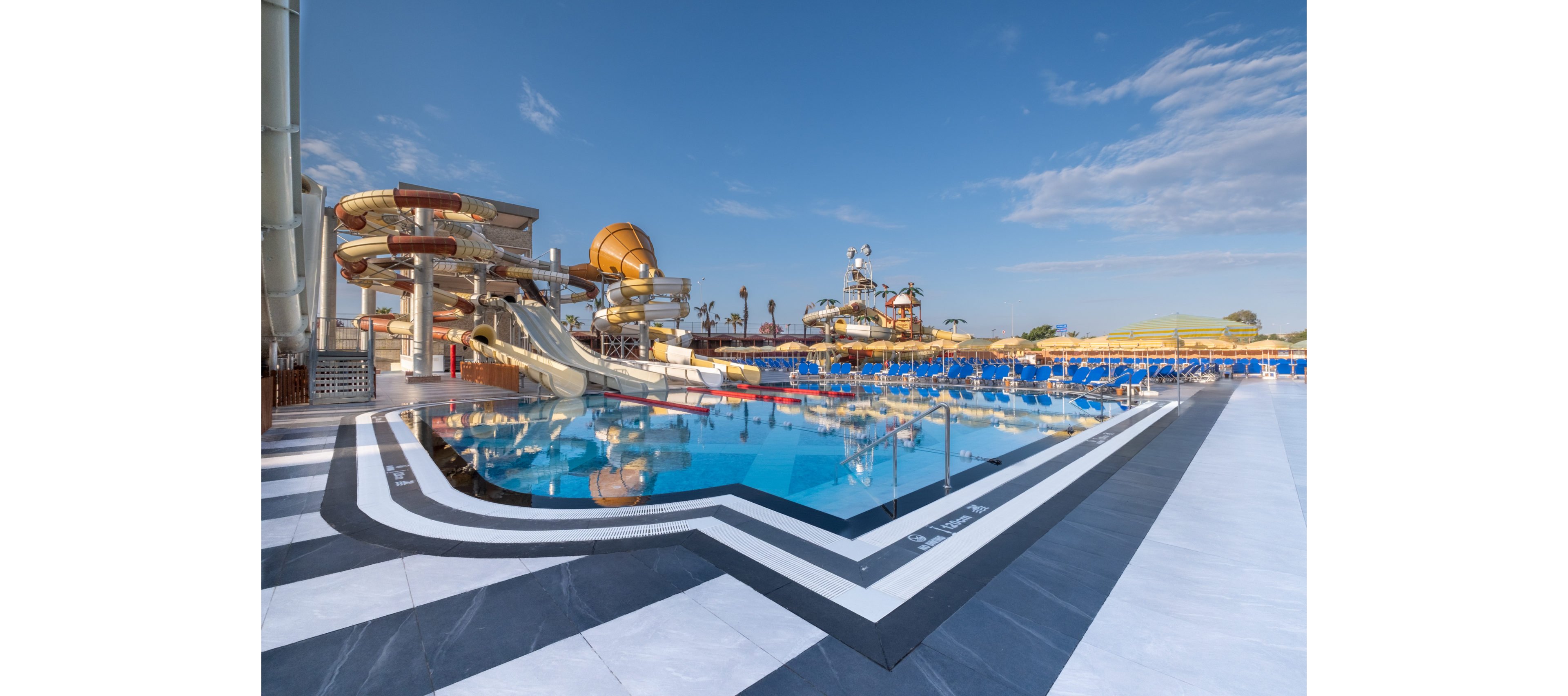 A large outdoor water park features a clear blue swimming pool in the foreground with geometric tile patterns around the deck. Multiple winding water slides and splash structures rise behind the pool, creating a playful and dynamic skyline. Rows of blue sun loungers and umbrellas line the far side of the pool, suggesting a resort setting for relaxation. The bright daylight and clear sky give the scene a warm, inviting vacation atmosphere.