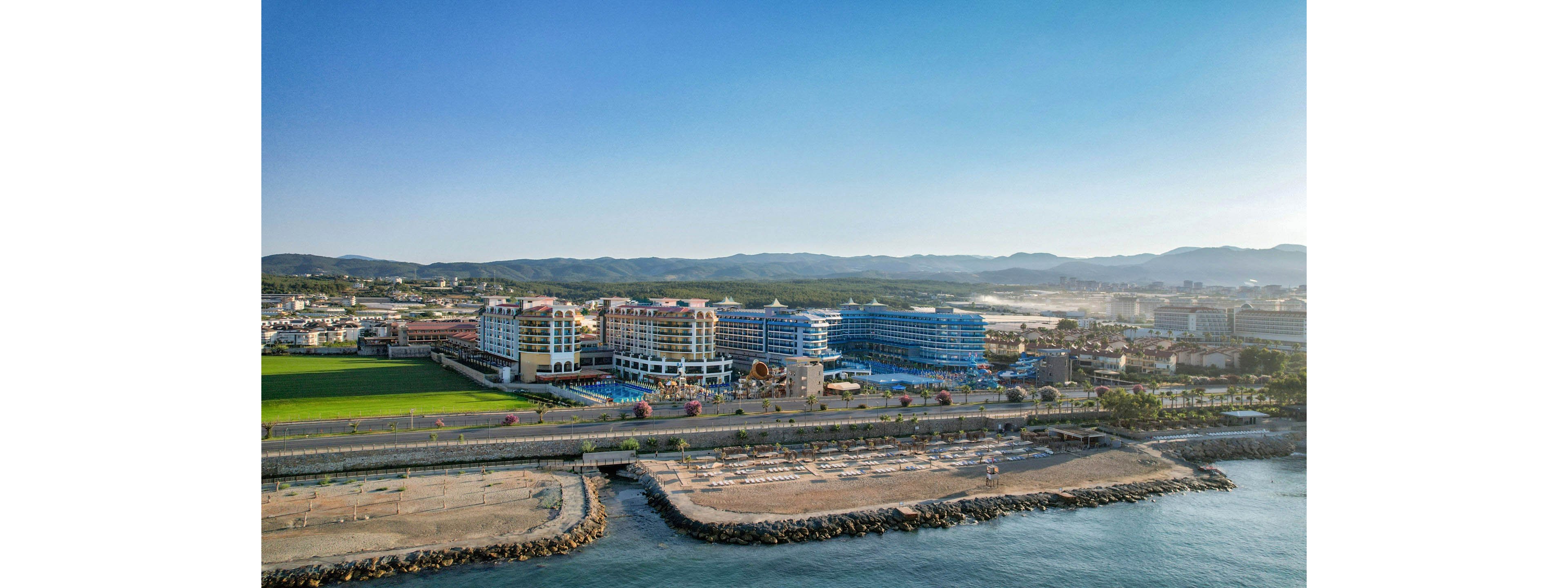 A large seaside resort complex stretches along the coastline, featuring multiple multi-story buildings with blue accents. In front of the hotels, a wide beachfront promenade and sandy beach area line the calm water. Sun loungers and umbrellas are arranged in rows on the sand, while a green sports field sits adjacent to the resort. The scene is captured from an elevated viewpoint under clear blue skies with distant hills in the background.