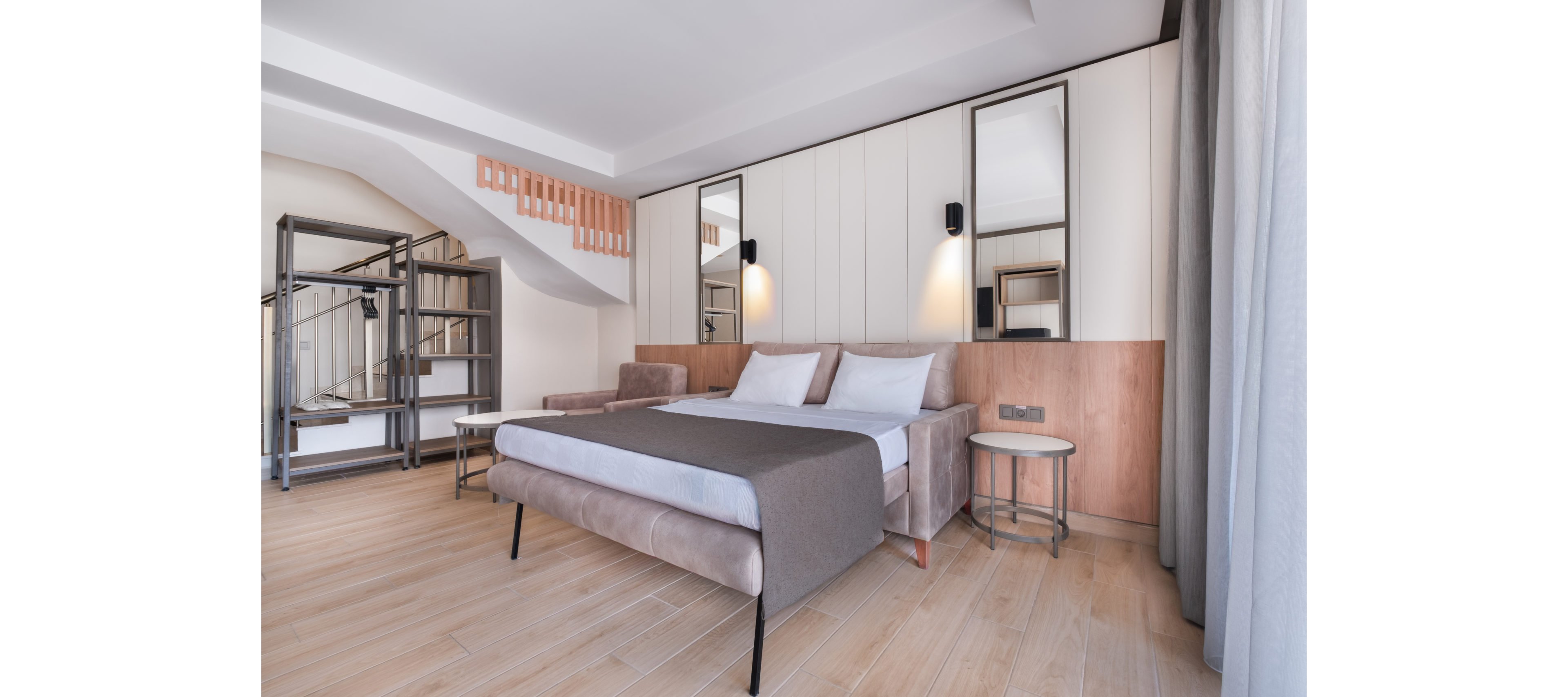 Contemporary hotel bedroom featuring a double bed with neutral bedding and upholstered frame. The room includes a sofa, side tables, and built-in shelving under a staircase. Light wood flooring and soft beige and gray tones create a clean, minimalist aesthetic. Large windows with curtains allow natural light to brighten the space, complemented by wall-mounted lamps and mirrors above the bed.