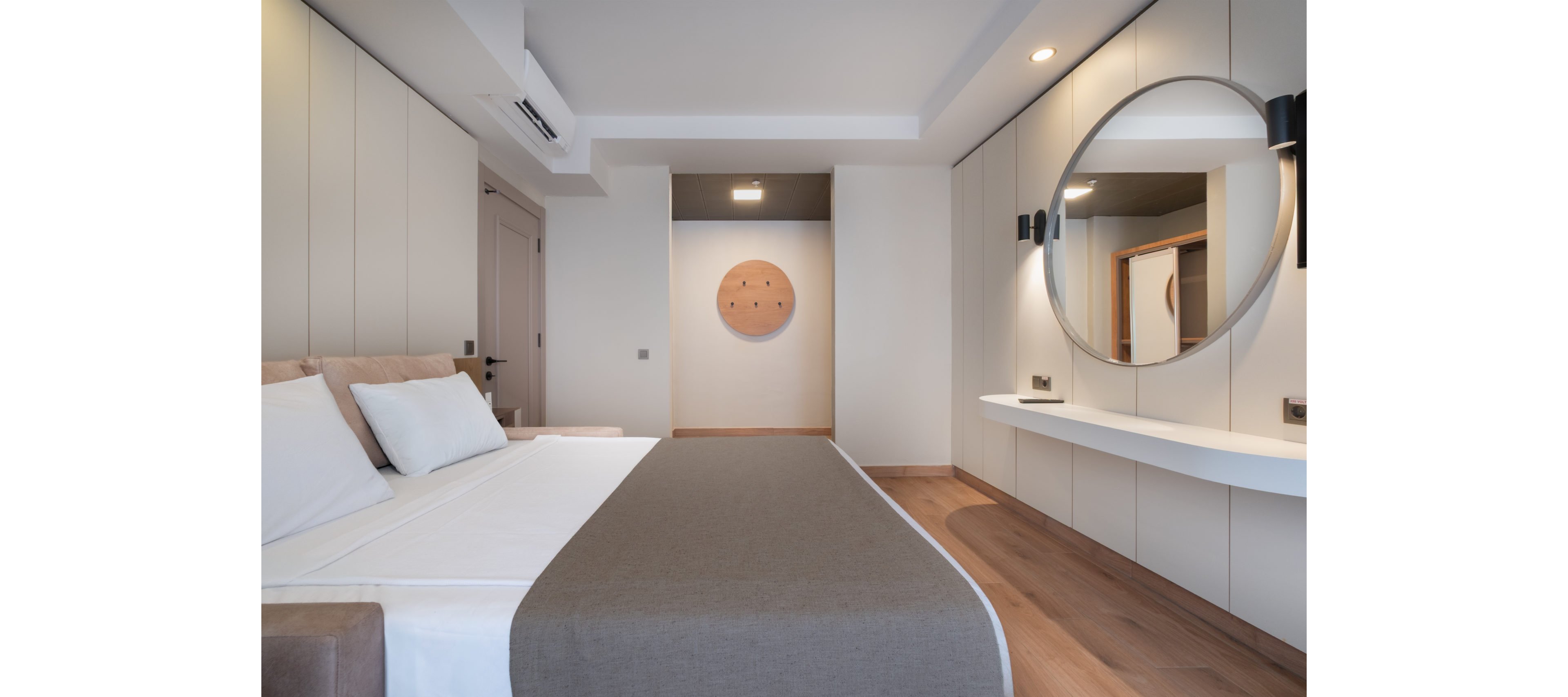 Contemporary bedroom with a neatly made double bed featuring white linens and a taupe bedspread. The room has built-in wall cabinets, a floating white console, and a large round mirror creating a clean, minimalist look. Warm wood flooring and soft neutral colors contribute to a calm, inviting atmosphere. A circular wall art piece and subtle recessed lighting add visual interest to the modern space.