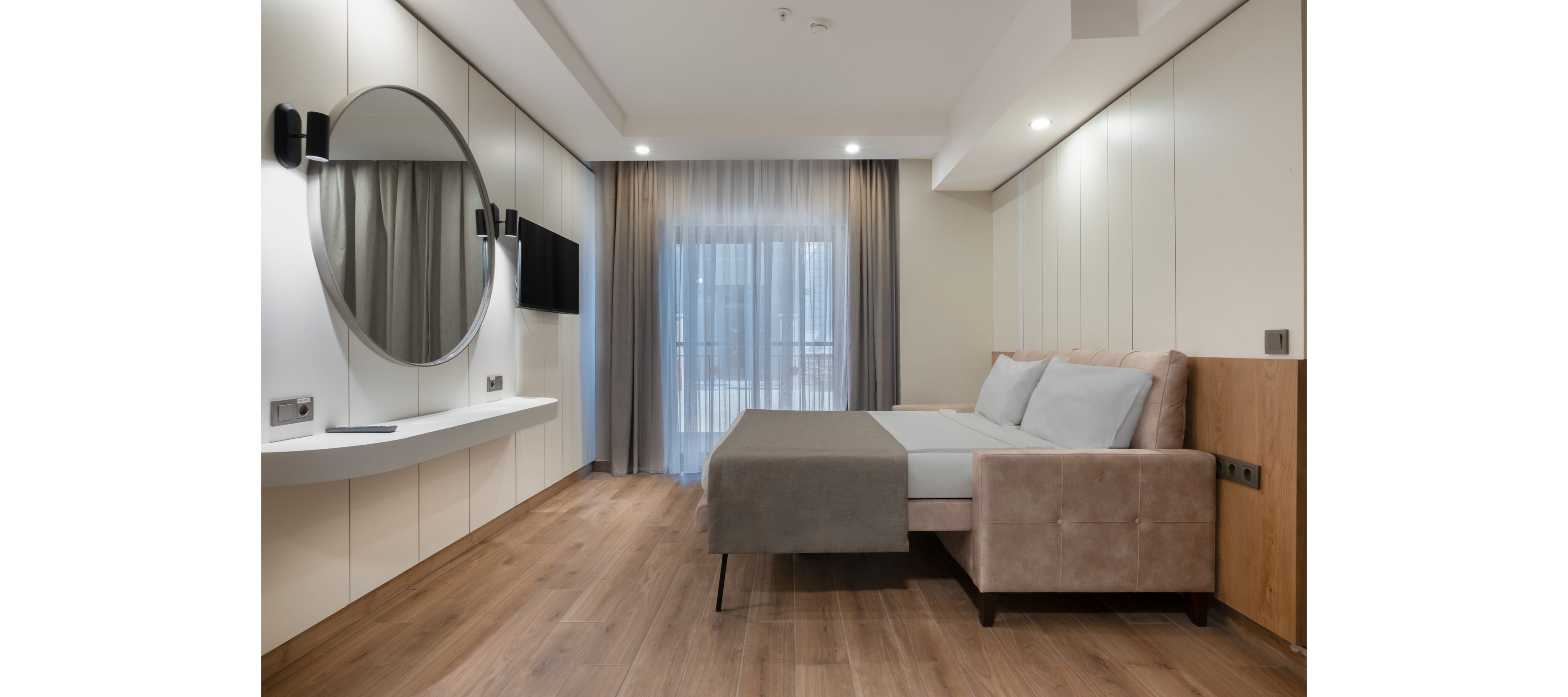 Contemporary bedroom interior featuring a beige upholstered bed with gray bedding. The room includes a wall-mounted flat-screen TV and a large round mirror above a sleek white console. Floor-to-ceiling curtains cover glass doors leading to a balcony, allowing natural light to enter. Warm wood flooring and neutral wall panels create a clean, minimalist aesthetic.
