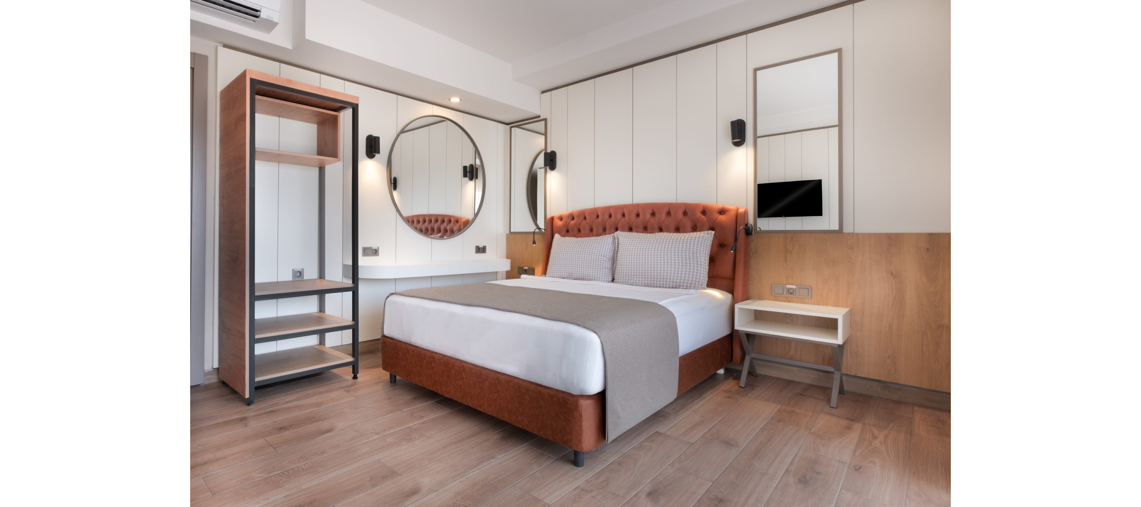 Contemporary hotel-style bedroom featuring a large upholstered bed with neutral bedding. The room includes a round wall mirror above a sleek white desk and an open shelving unit to the left. Warm wood paneling, light-colored walls, and minimalist bedside tables create a clean, inviting atmosphere. Soft lighting and simple decor emphasize a modern, comfortable design.