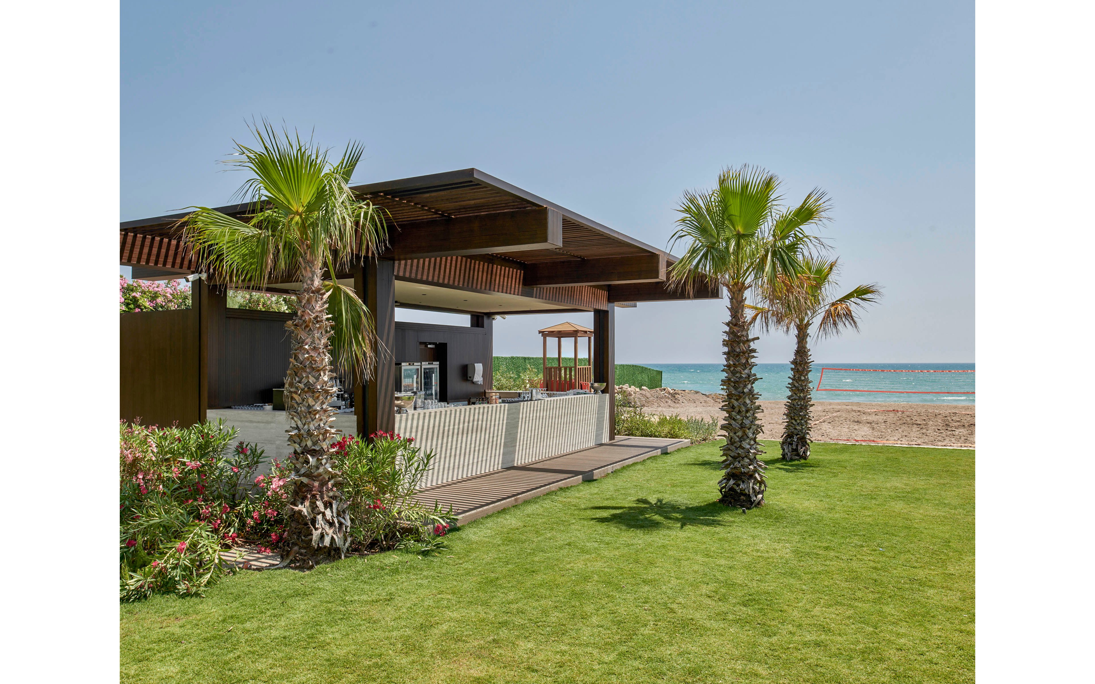 A contemporary open-air pavilion stands on a manicured lawn beside a sandy beach. Palm trees and flowering shrubs surround the wooden structure, creating a lush tropical atmosphere. The calm blue sea and clear sky form a serene backdrop, suggesting a relaxing coastal retreat. Clean lines and natural materials emphasize a modern, minimalist outdoor design.