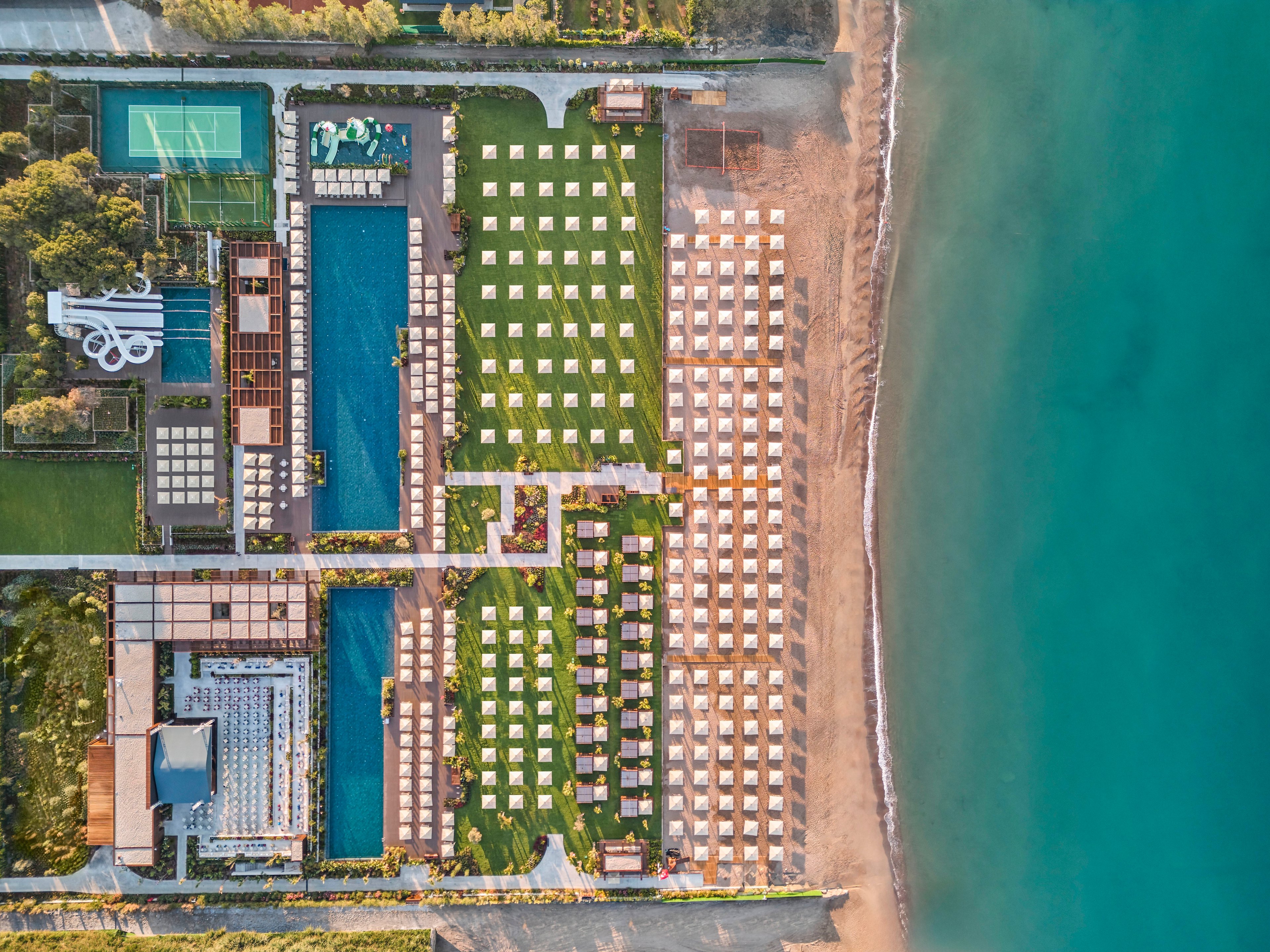 A large seaside resort complex is captured from above, showing orderly rows of sun loungers and umbrellas along the beach. Multiple rectangular swimming pools and green lawns are arranged in a geometric layout beside the shoreline. Recreational areas, including a tennis court and waterslide structure, are visible among landscaped gardens and pathways. The turquoise sea borders the right side of the image, contrasting with the warm tones of sand and the resort’s architecture.