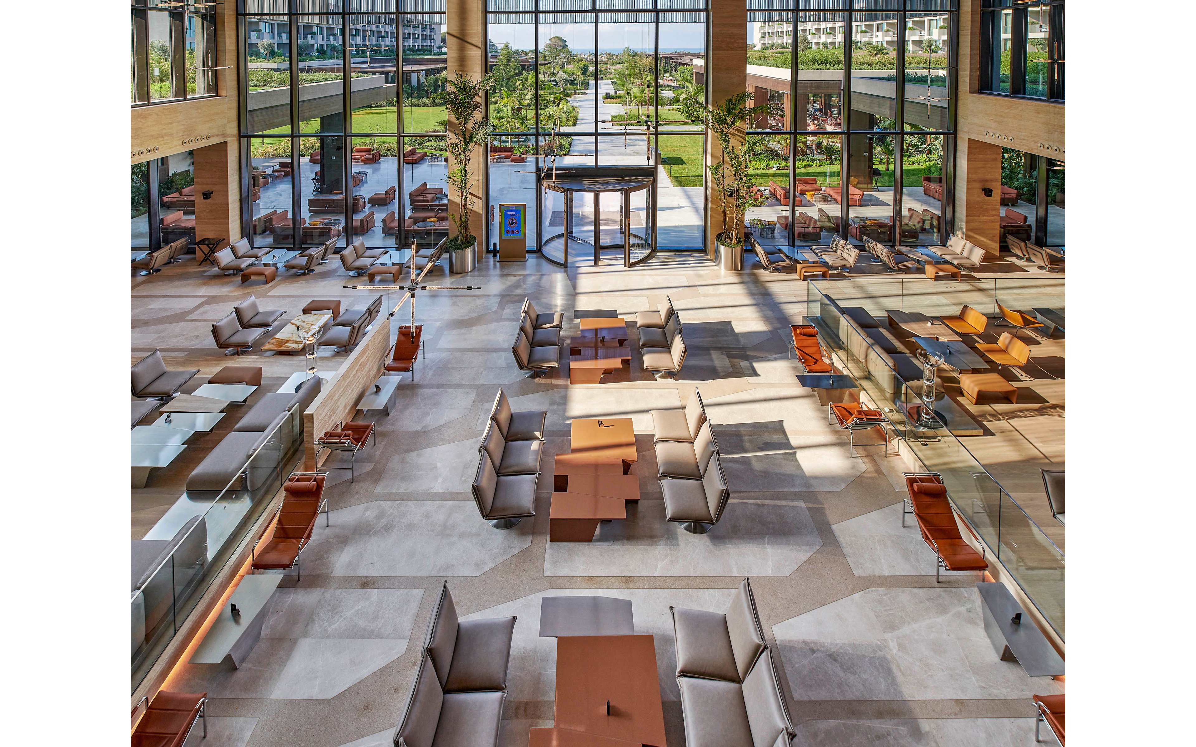 A large contemporary lobby features multiple seating areas with sofas and armchairs arranged around low tables. Floor-to-ceiling windows open to a landscaped outdoor terrace and green lawn beyond. Warm wood accents, neutral upholstery, and glass railings create a clean, upscale atmosphere. The space appears quiet and empty, emphasizing openness and architectural design.