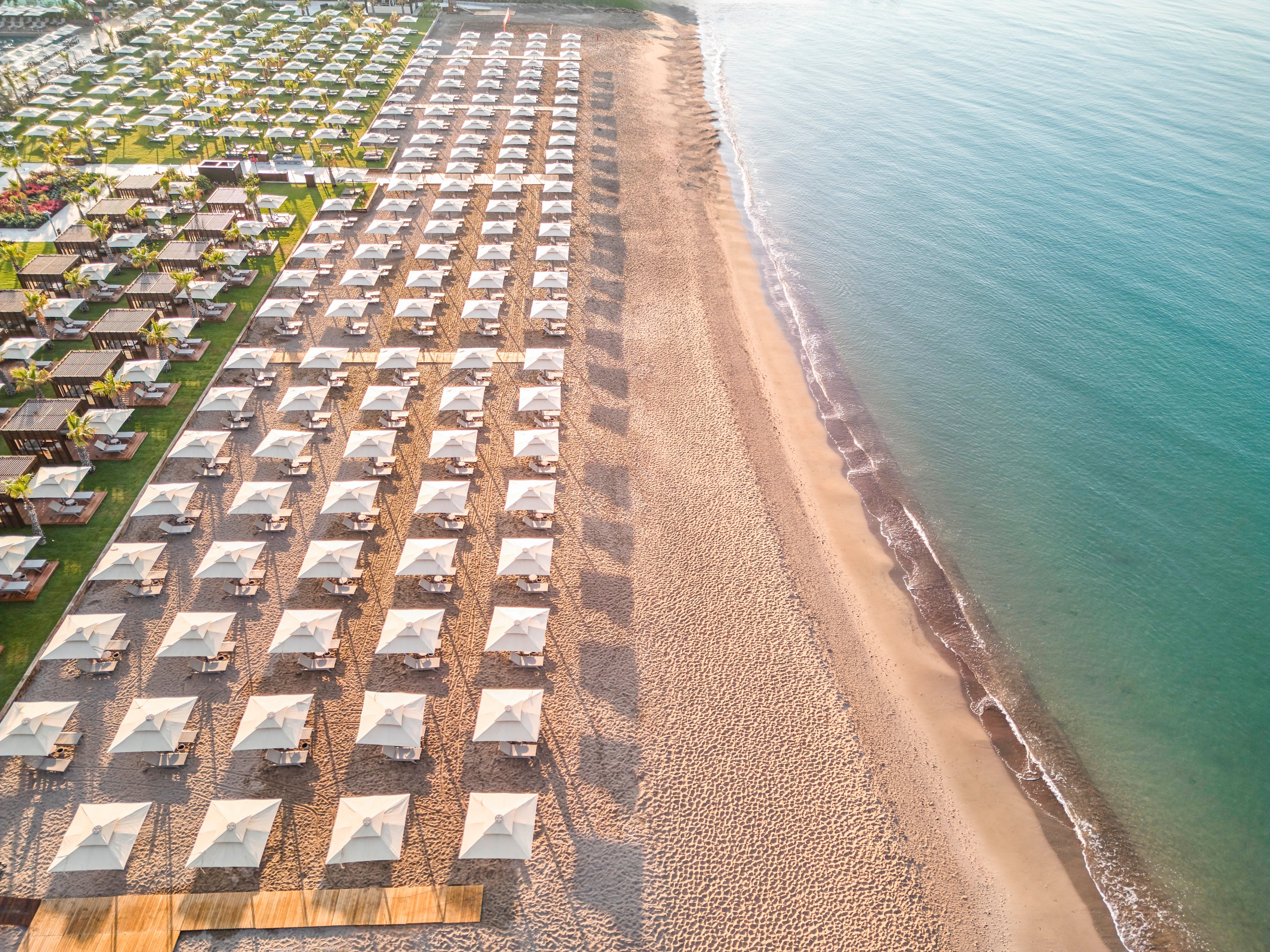 A drone captures an expansive beachfront lined with neatly arranged white cabanas. The cabanas are set in precise rows on sand and adjacent green lawn, creating a strong geometric pattern. Gentle waves meet the shoreline on the right side, adding a calm coastal element. The warm, soft lighting suggests a tranquil sunrise or sunset atmosphere.