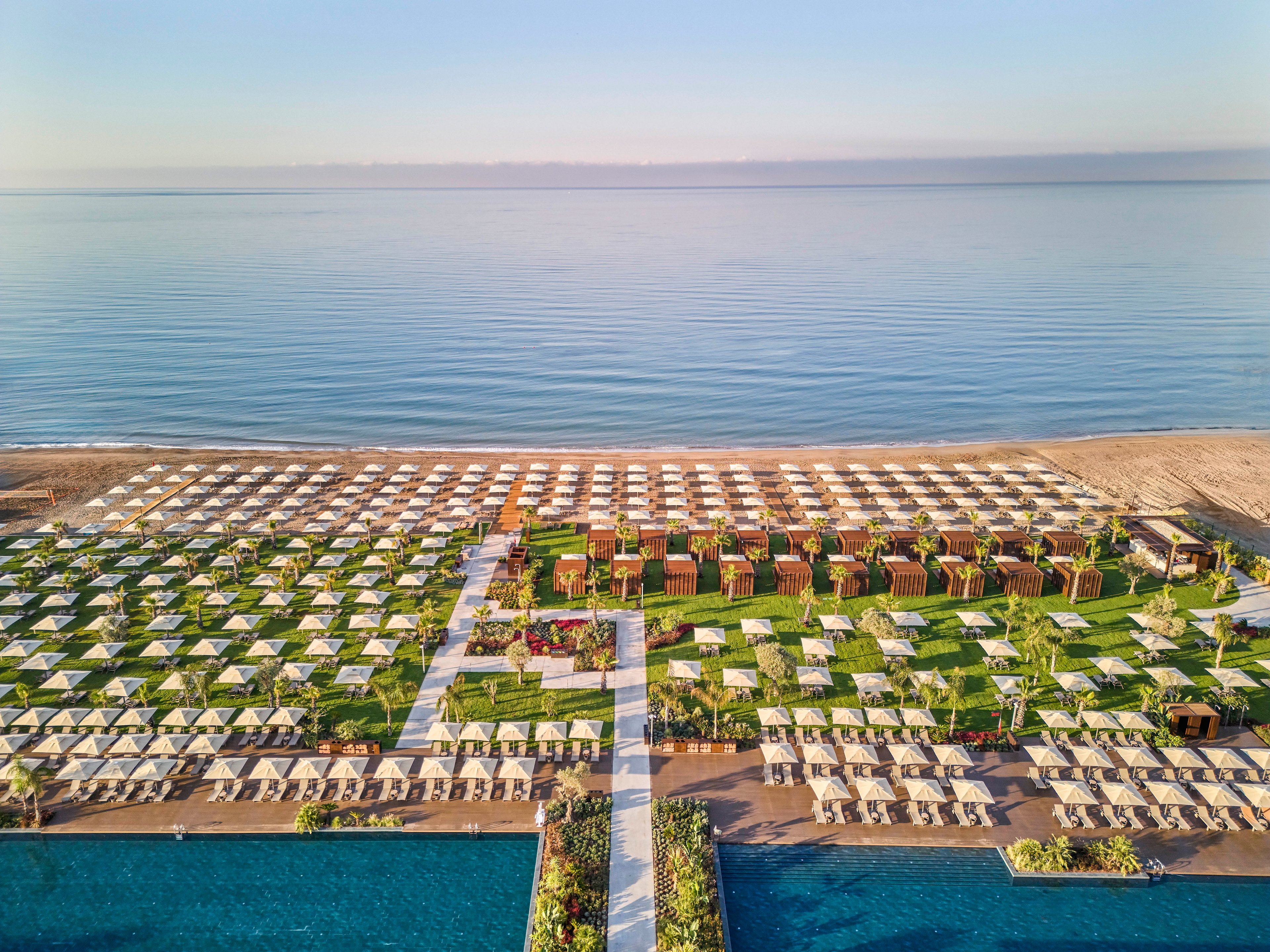 A large beachfront resort is captured from above, showing rows of sun loungers and umbrellas arranged in a precise grid. Green lawns with pathways and cabanas separate the seating areas, leading toward a sandy beach and calm sea. Two blue swimming pools frame the foreground, contrasting with the warm tones of the sand and the soft pastel sky. The overall scene feels tranquil, orderly, and inviting for vacation relaxation.
