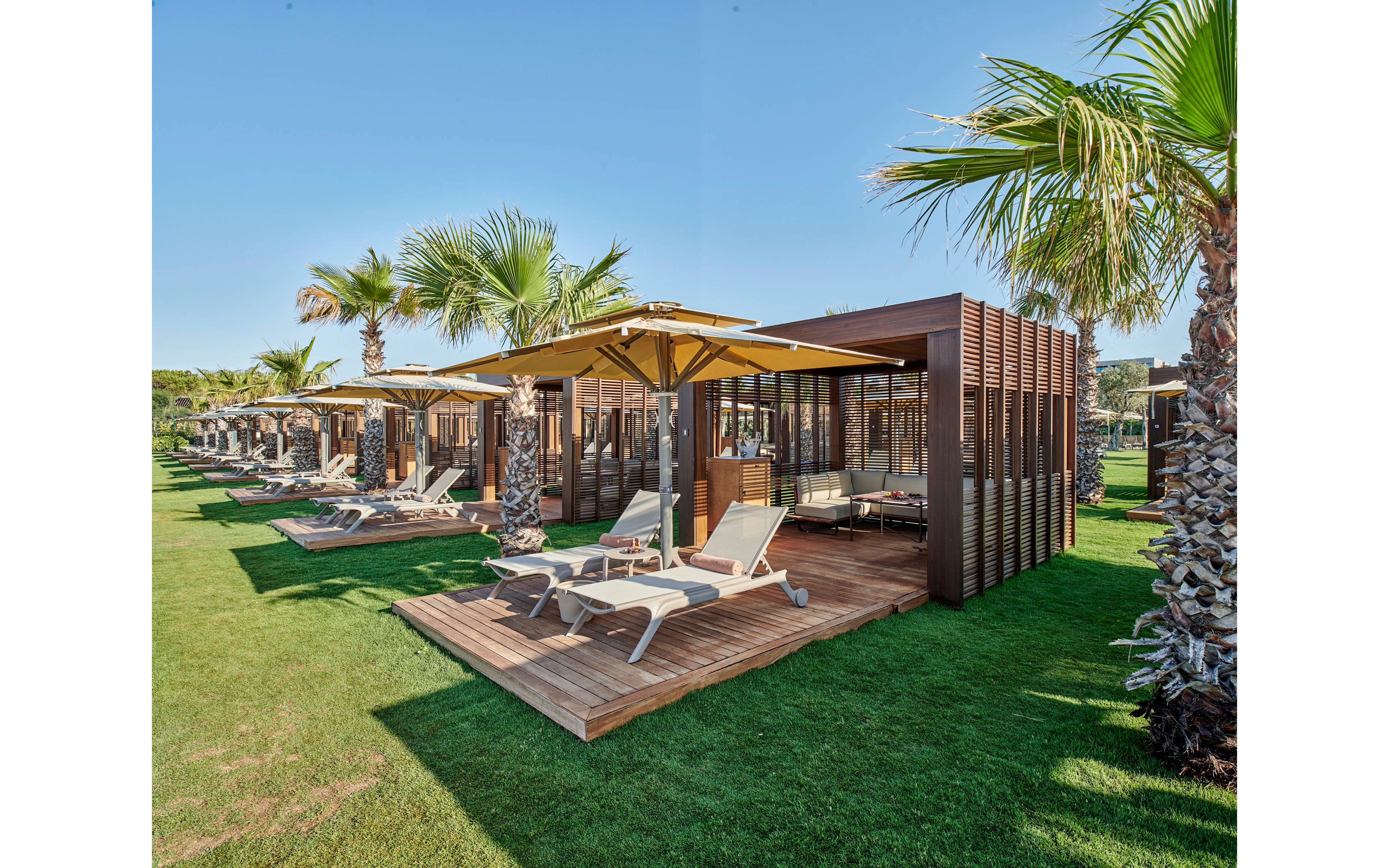 A row of contemporary wooden cabanas with yellow sun umbrellas stands on a neatly manicured lawn. Each cabana features white sun loungers and shaded seating areas, creating a relaxed resort atmosphere. Palm trees line the space, adding a tropical feel under clear blue skies. The scene suggests a luxurious outdoor retreat near the coast.