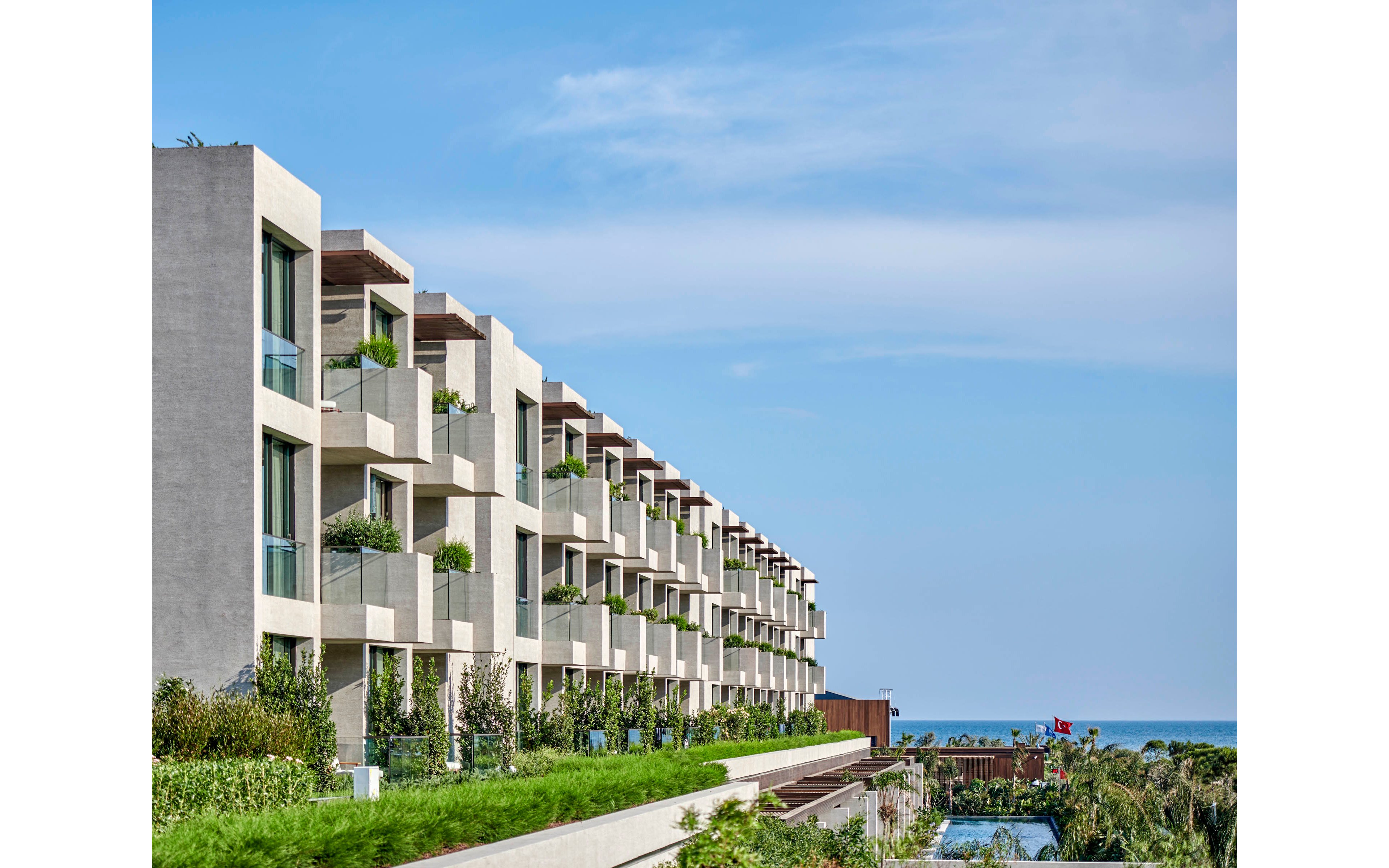 A contemporary multi-story apartment building features rows of balconies facing the sea. Lush greenery and landscaped terraces line the lower levels, adding a natural contrast to the clean architectural lines. The scene is set under a clear blue sky with the ocean visible in the background. The overall atmosphere is calm, bright, and inviting, suggesting a coastal residential or resort setting.