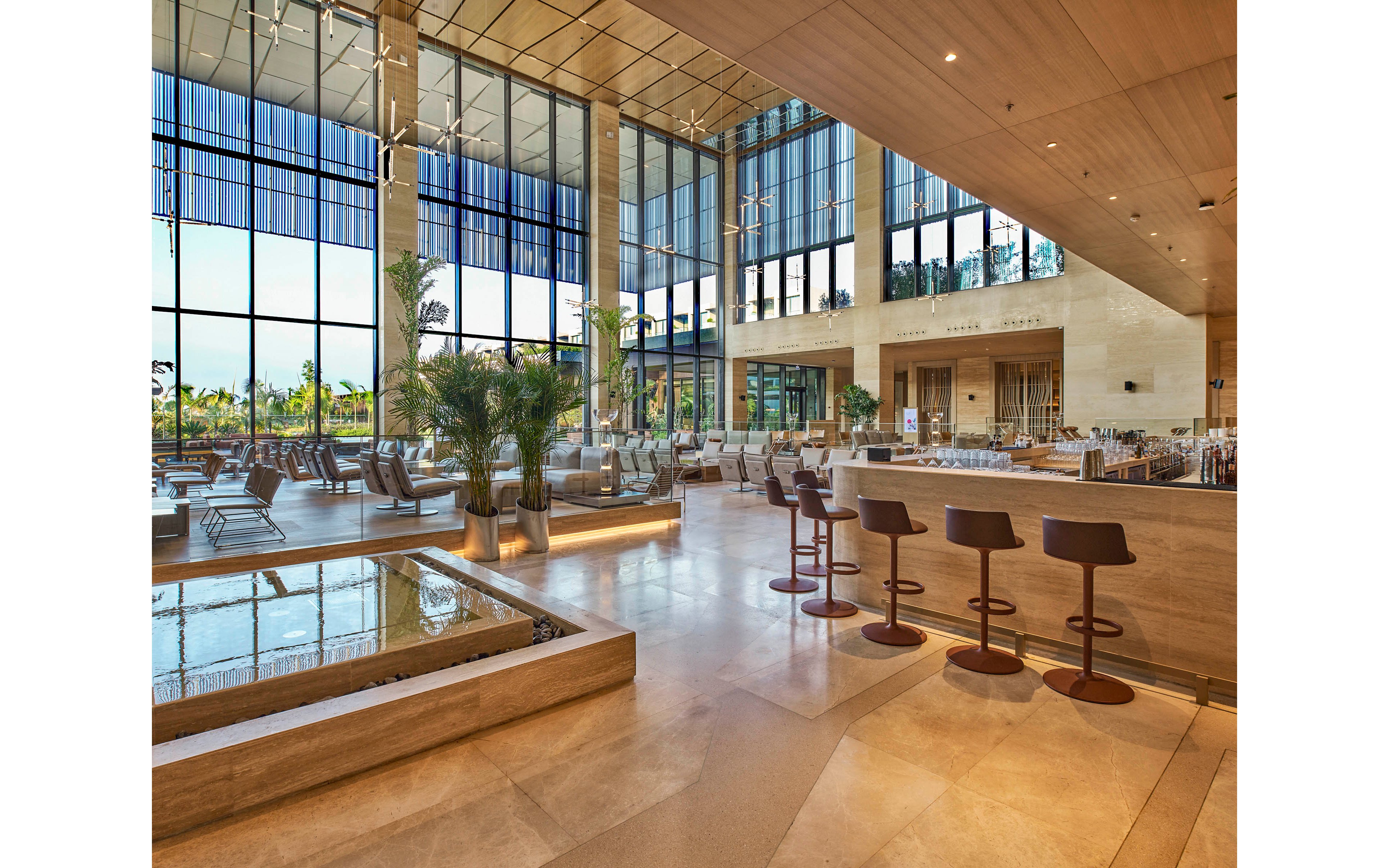 Large contemporary lobby features a sleek bar area with high stools and polished stone flooring. Floor-to-ceiling windows flood the space with natural light and offer views of greenery outside. Indoor palm plants, water feature, and neutral beige tones create a luxurious, resort-like atmosphere. Minimalist furniture and clean architectural lines emphasize an upscale, modern design.
