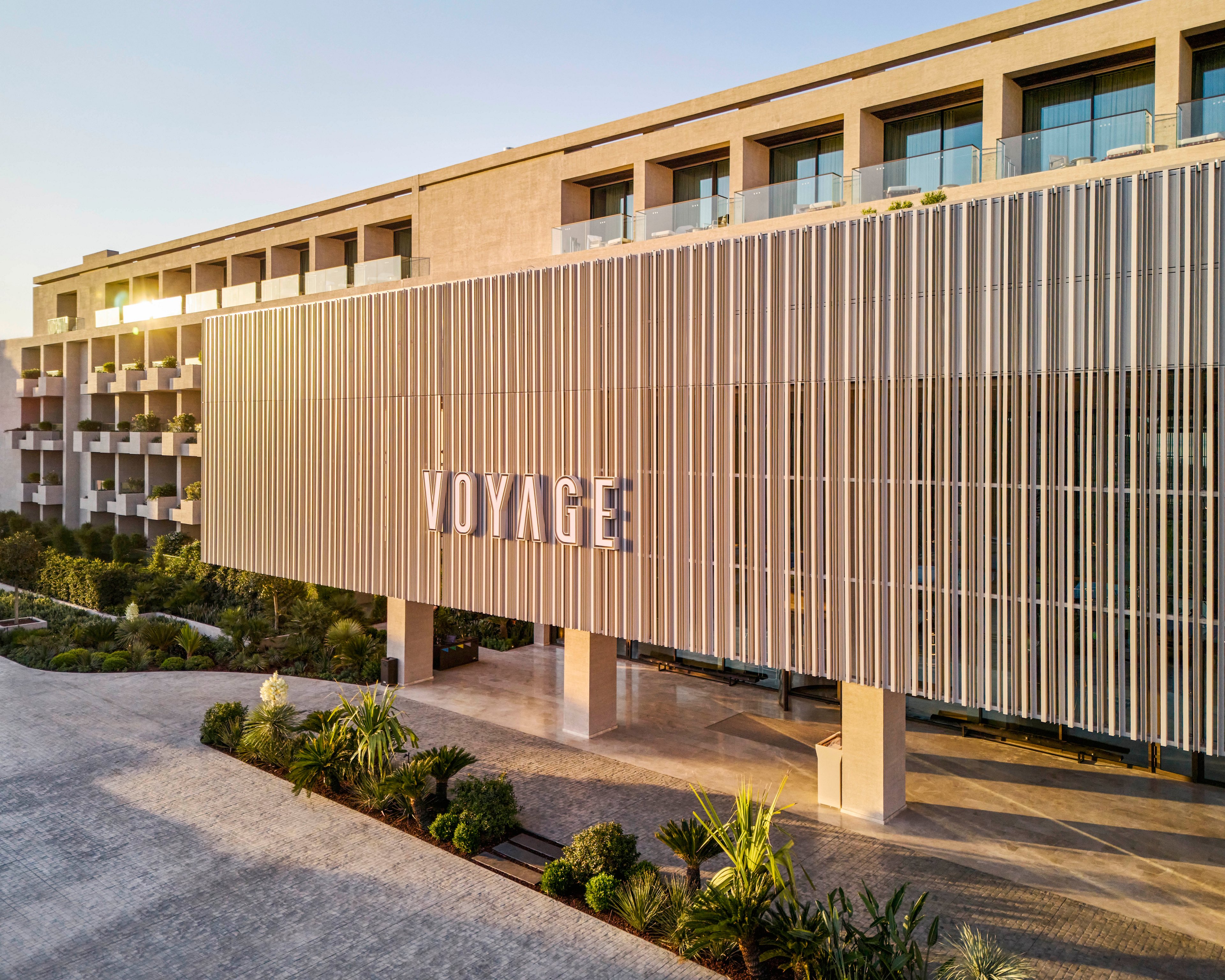 A contemporary multi-story hotel building features a prominent facade with the word VOYAGE displayed in large metallic letters. Vertical slats create a sleek architectural screen across the front entrance area. The scene is captured in warm early morning or late afternoon light, highlighting clean lines and neutral tones. Lush green landscaping and a paved driveway frame the modern resort-style property.
