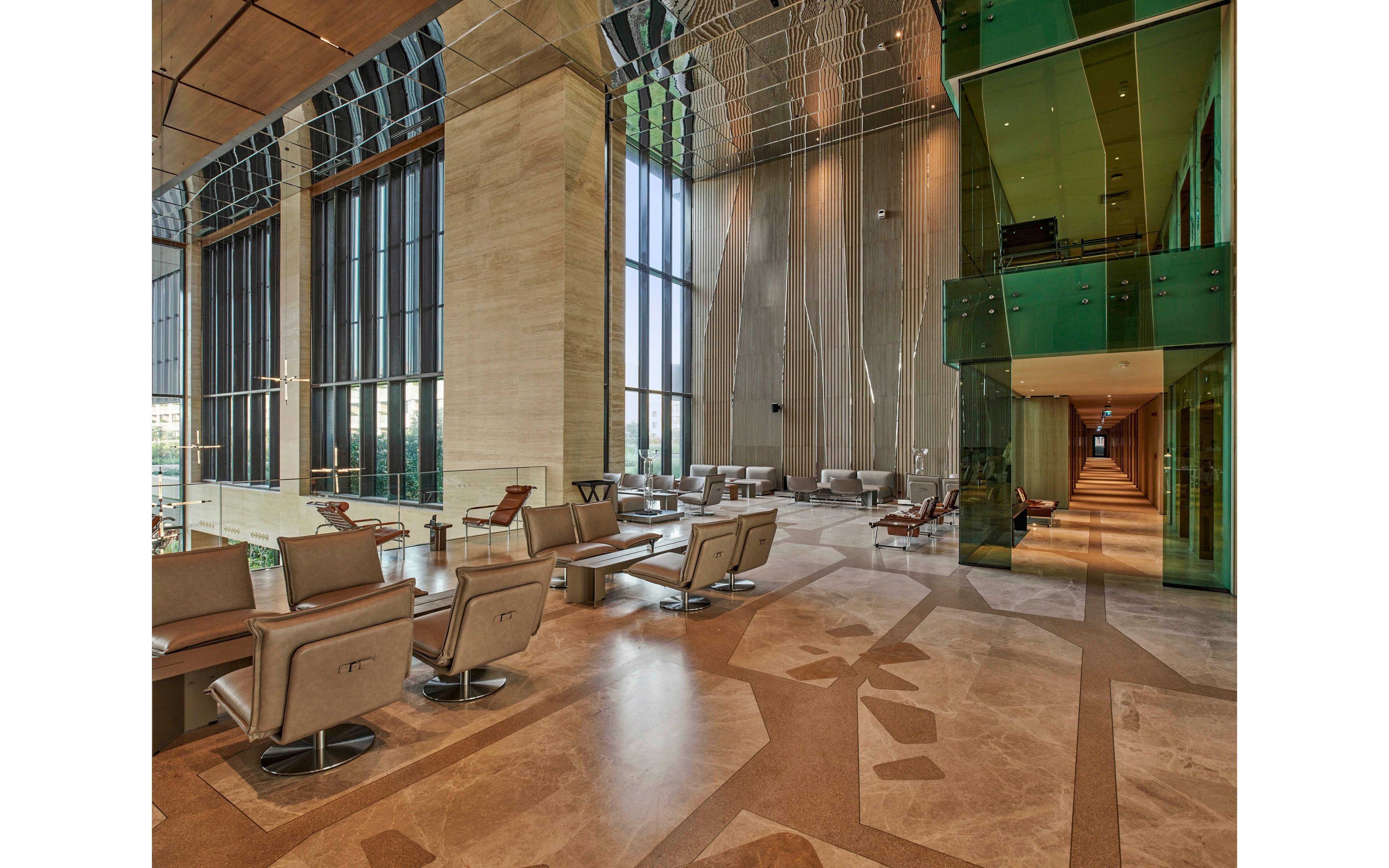 A large contemporary lobby features comfortable lounge chairs and small tables arranged near tall floor-to-ceiling windows. Natural light floods the space, highlighting the polished stone floors and high reflective ceiling with decorative elements. A green glass-walled corridor extends along one side, adding a bold architectural accent. The overall atmosphere is open, luxurious, and designed for relaxed waiting or informal meetings.