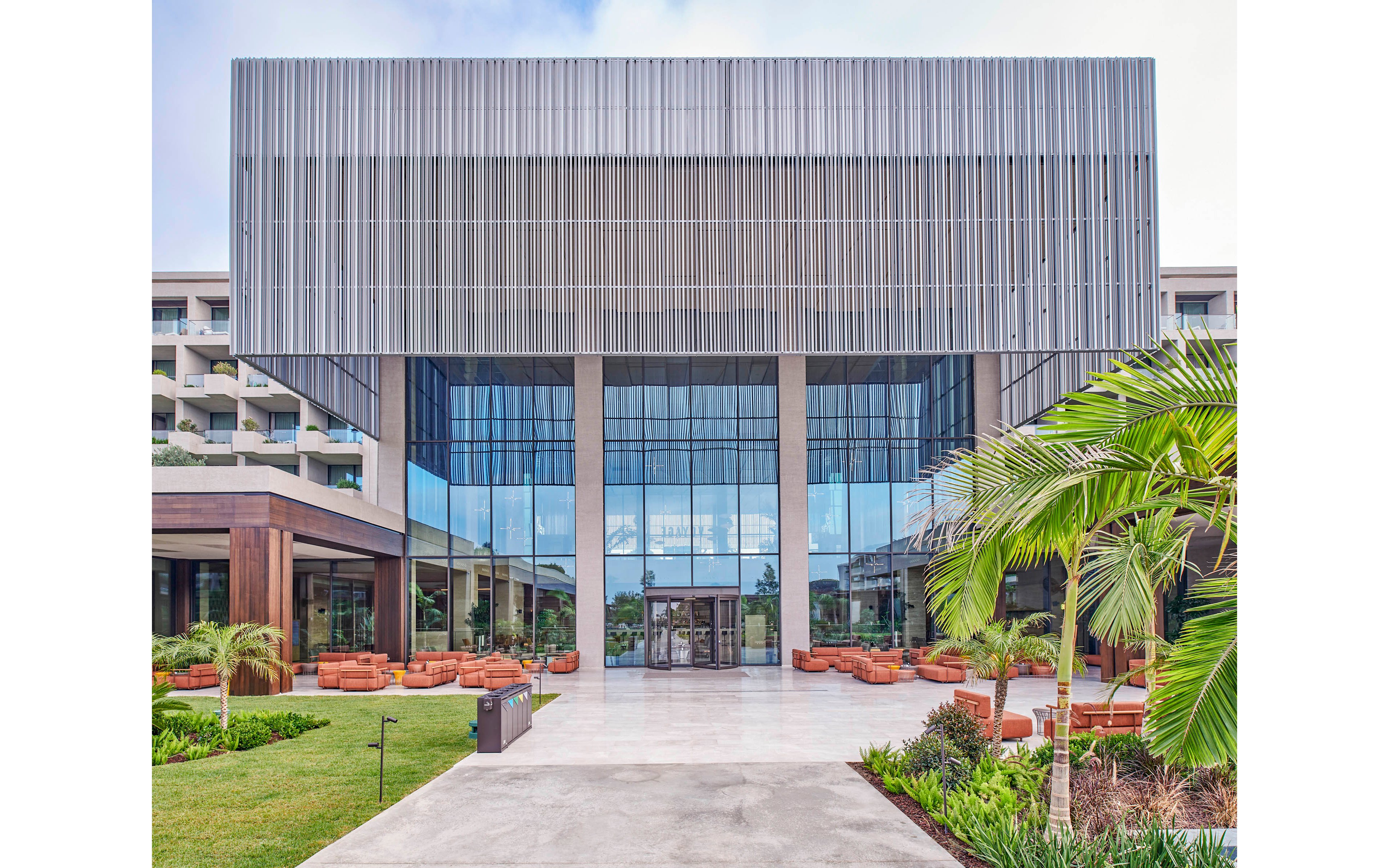 A contemporary resort building features a tall glass entrance framed by vertical metal slats. The foreground shows a wide concrete walkway leading to the main doors, bordered by manicured green lawns and tropical plants. Outdoor seating areas with cushioned chairs are arranged on both sides of the entrance. The overall scene conveys a clean, modern architectural style in a bright daytime setting.