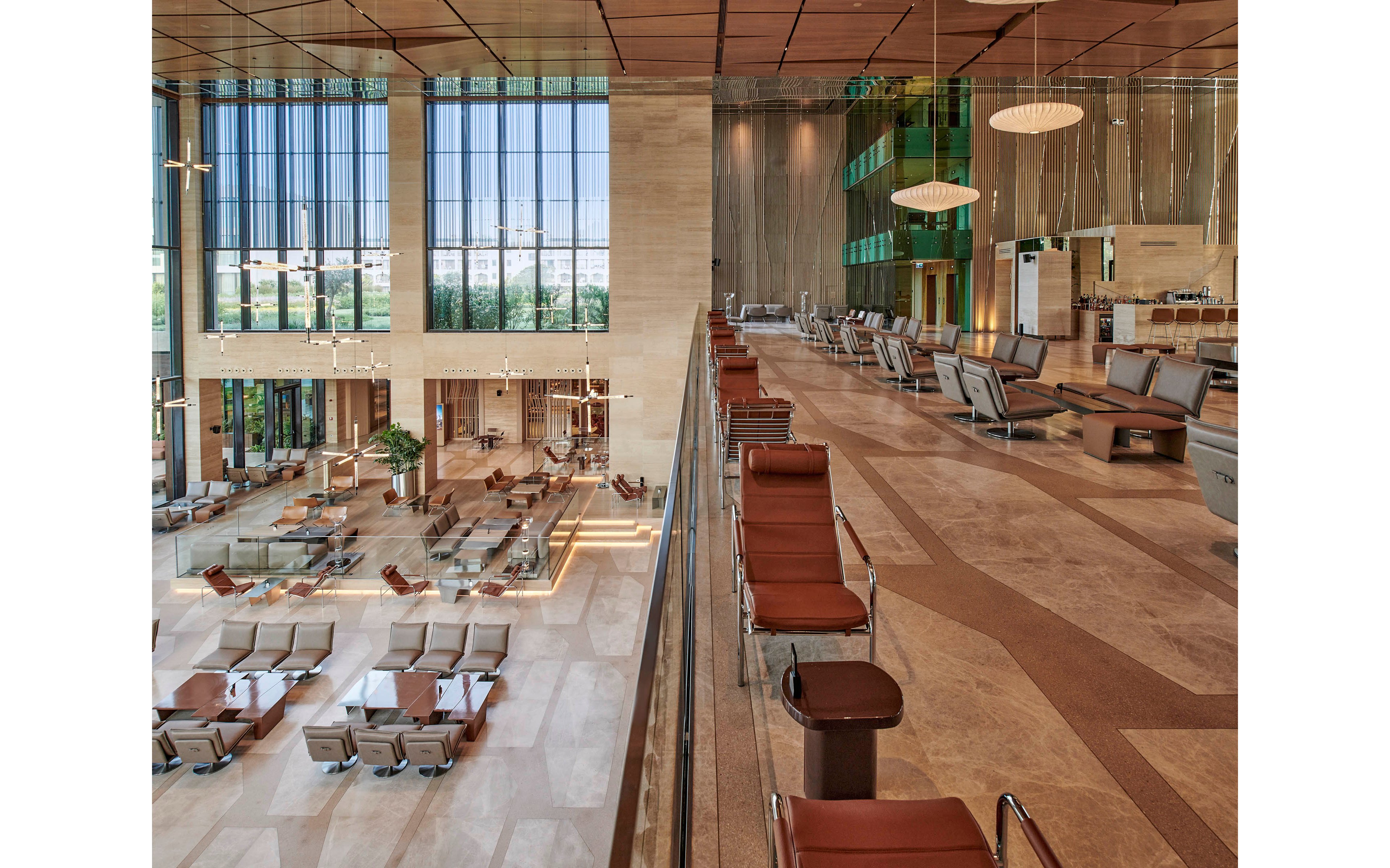 Large contemporary interior lobby features expansive seating areas and high ceilings. Floor-to-ceiling windows on one side let in abundant natural light and reveal greenery outside. Warm beige and brown tones dominate the marble floors, walls, and leather chairs. The space includes multiple tables, lounge chairs, and elevated seating zones, creating an open, luxurious atmosphere.