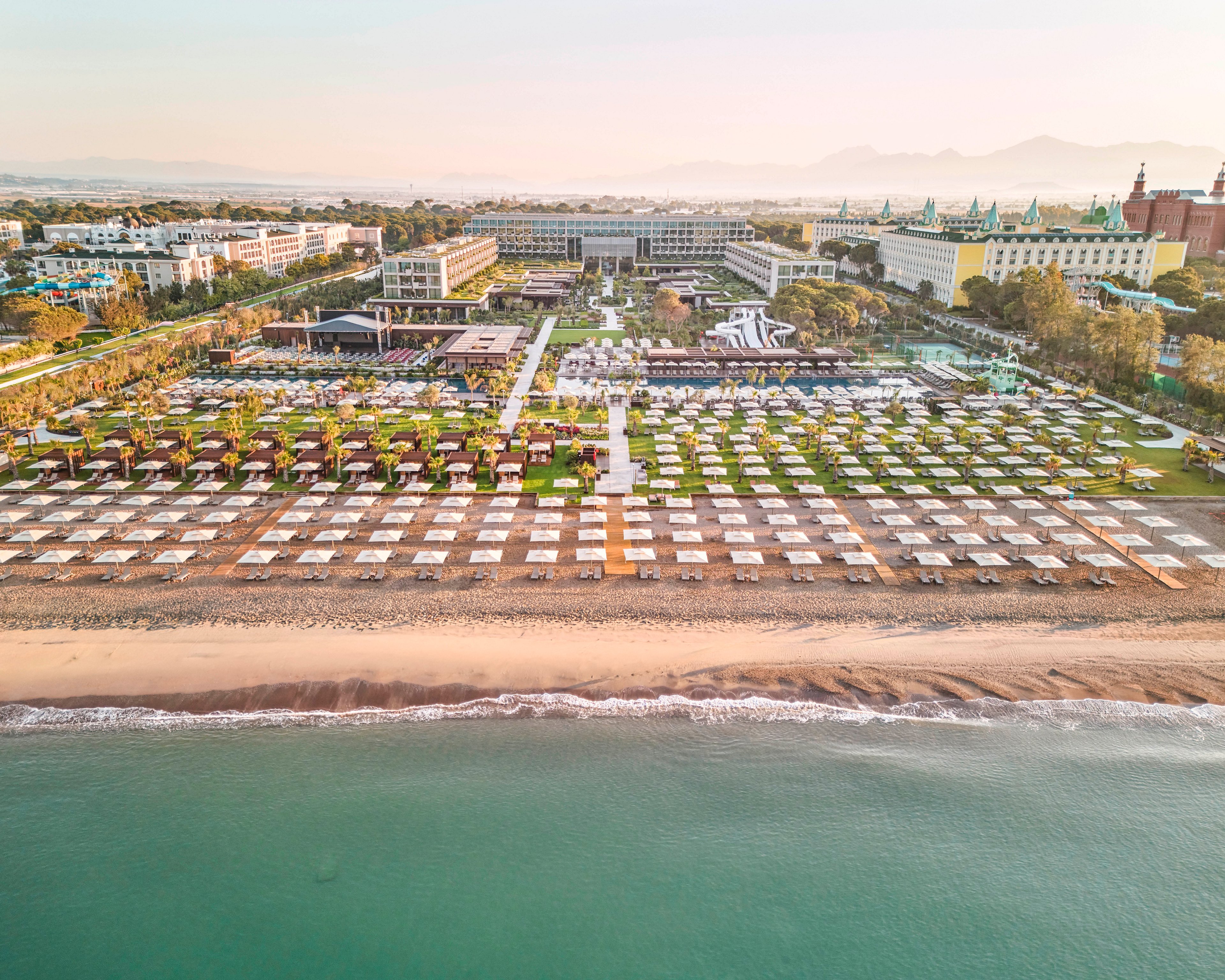 A large seaside resort complex stretches along a sandy beach beside calm turquoise water. Neatly arranged rows of sun loungers and umbrellas line the shore, leading back to pools, gardens, and multi-story hotel buildings. Palm trees and landscaped green areas create a structured, geometric pattern across the property. Soft sunrise or sunset light casts a warm glow over the scene, with distant mountains faintly visible on the horizon.