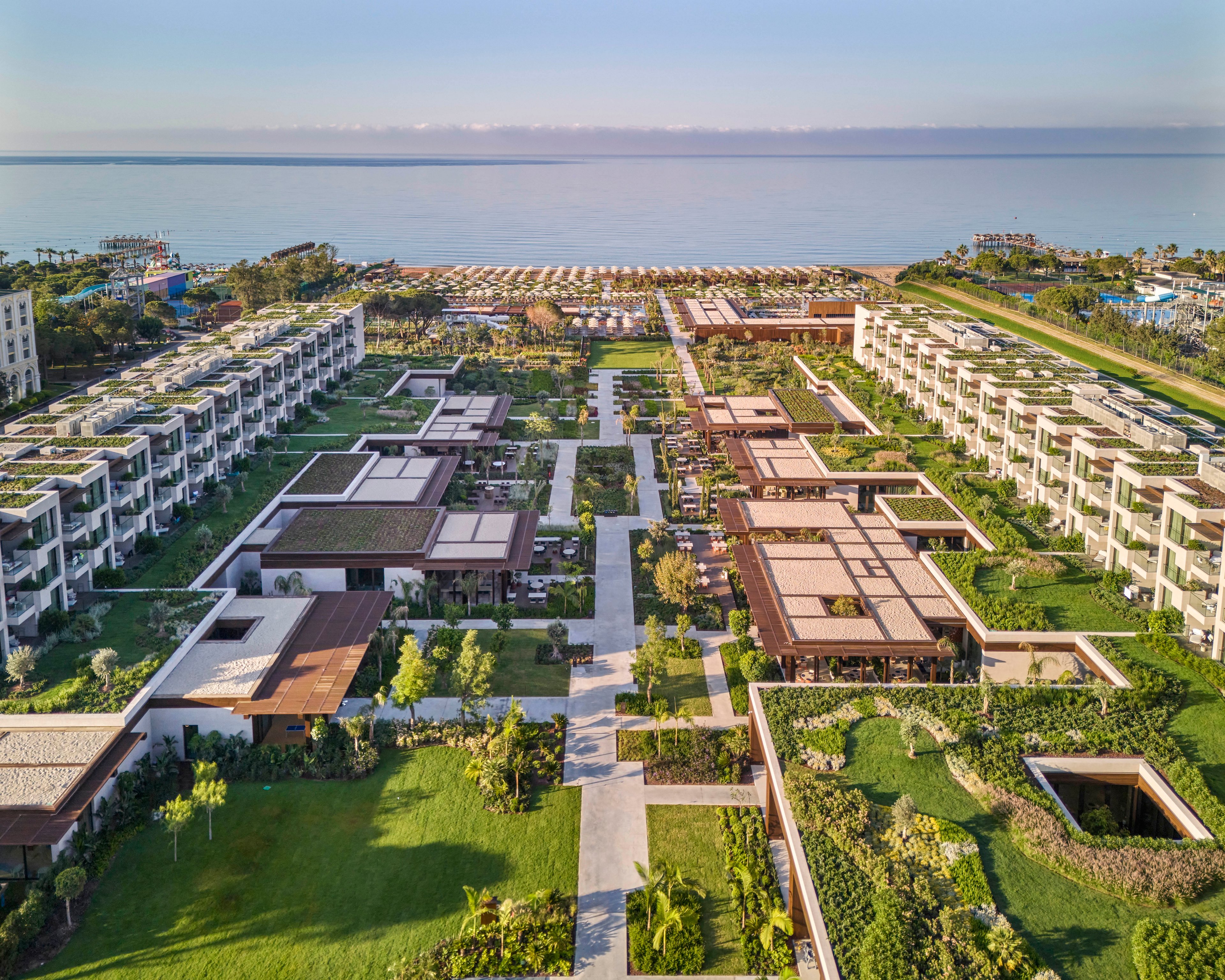 A large contemporary resort complex is captured from above, stretching toward a calm beachfront and open sea. Symmetrical rows of low-rise buildings with flat roofs frame landscaped courtyards and lush green gardens. Pathways and lawns create a structured geometric layout, with pools and terraces integrated into the design. In the distance, beach umbrellas and coastal vegetation line the shore under a clear sky.