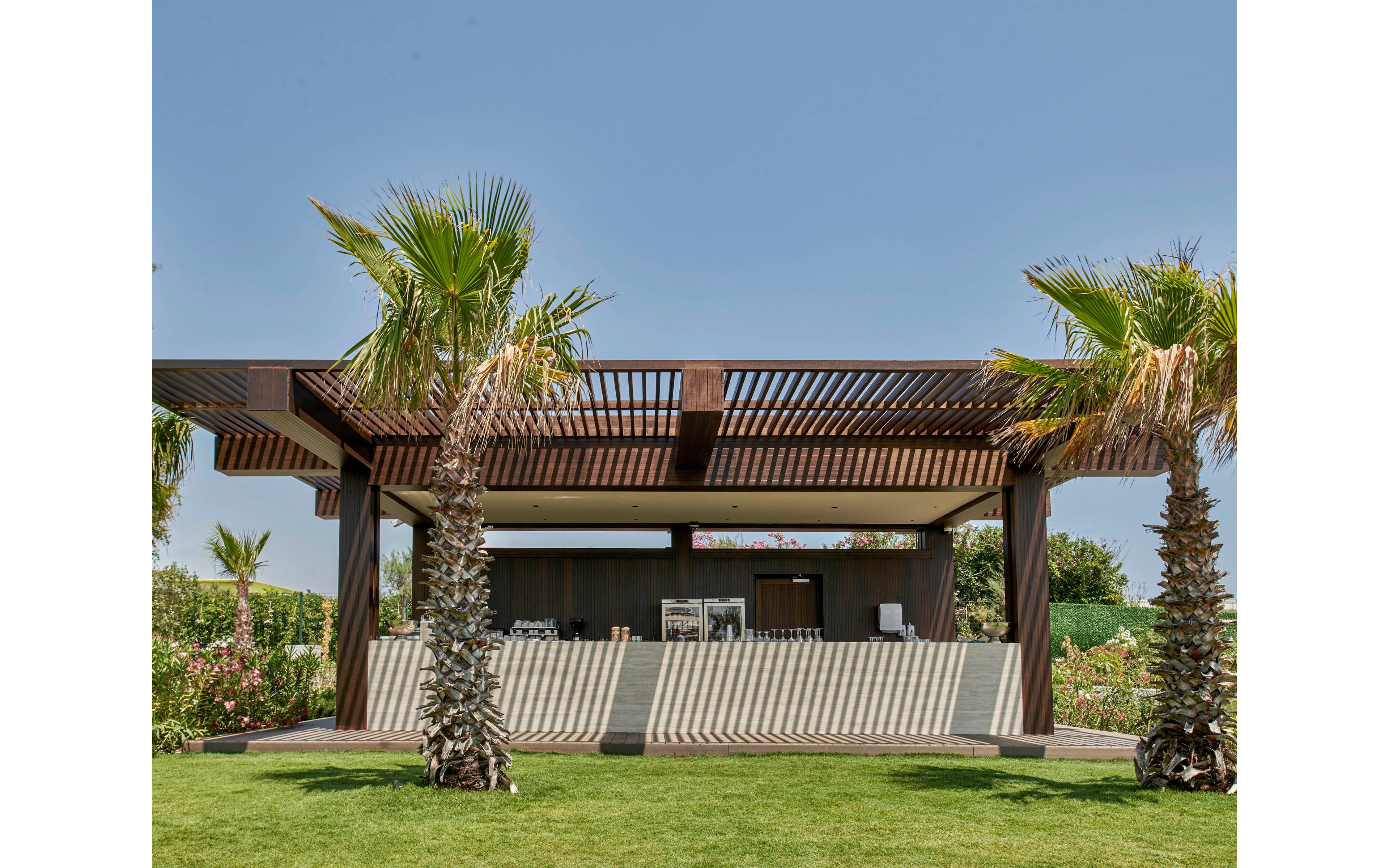 A contemporary open-air pavilion stands in a landscaped garden under a clear blue sky. The structure features wooden slats creating linear shadows across the light-colored base. Two tall palm trees frame the pavilion, with lush greenery and flowering shrubs surrounding the area. The scene suggests a relaxed outdoor leisure or hospitality setting.