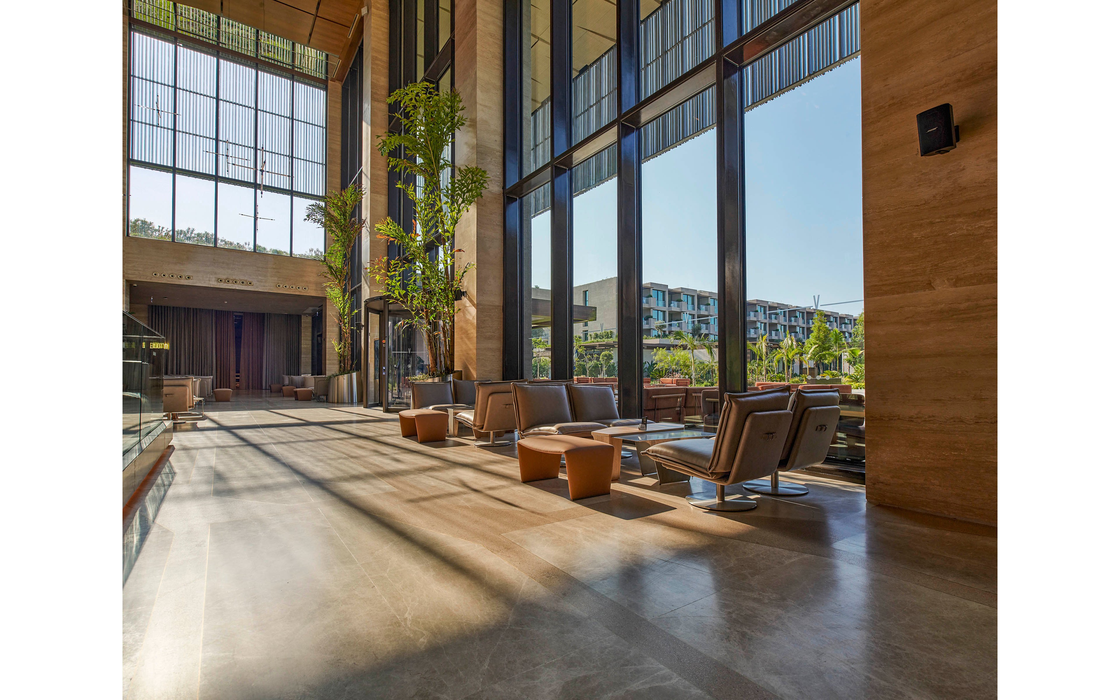 A spacious contemporary lobby features comfortable lounge chairs and low tables arranged along tall glass windows. Warm stone flooring and wall panels create a refined, upscale atmosphere. Sunlight streams through the floor-to-ceiling windows, casting long shadows and revealing a view of modern residential buildings outside. Tall indoor plants add a touch of greenery to the clean, minimalist interior design.