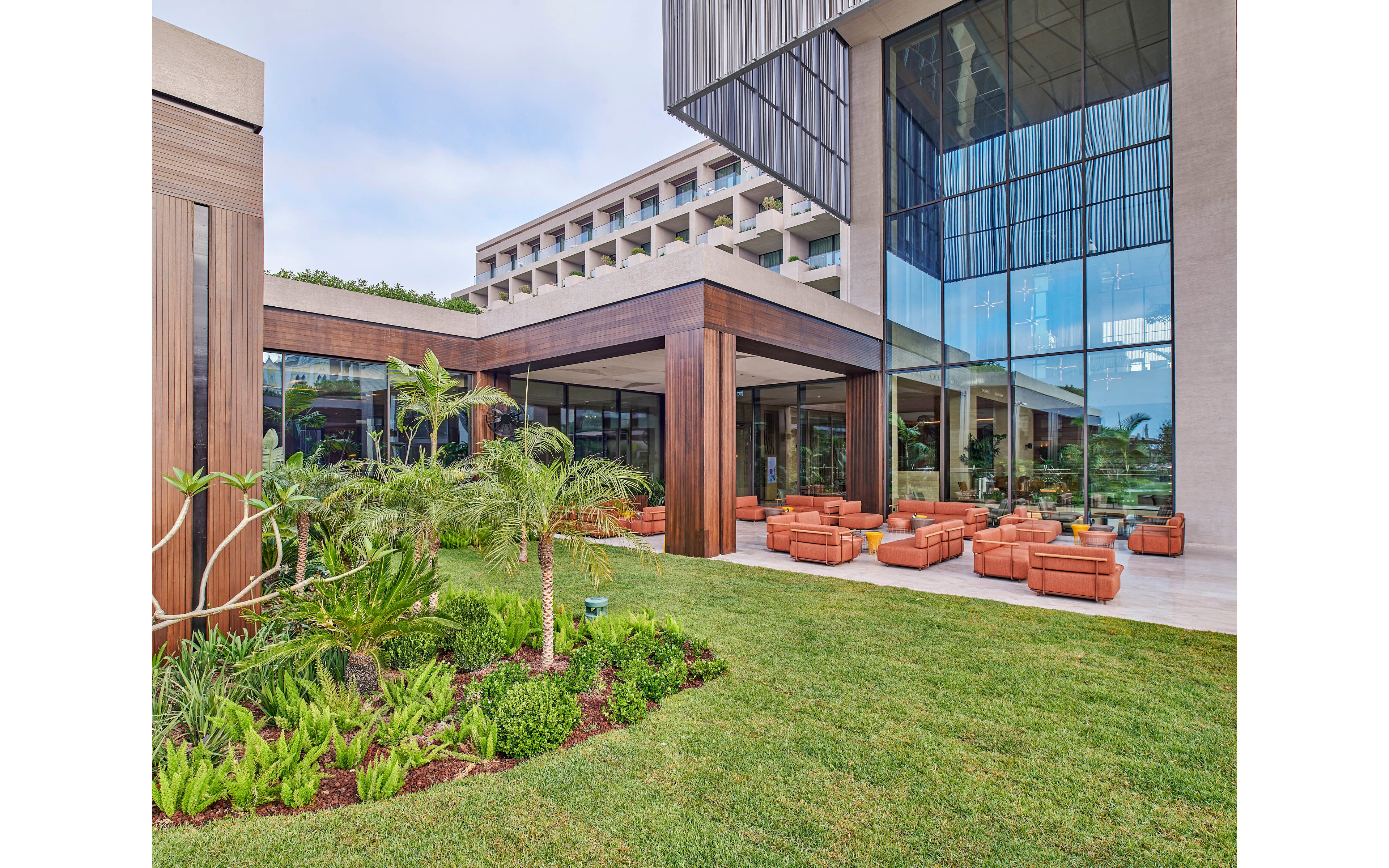 A contemporary resort or hotel building features a spacious courtyard with large glass windows and geometric architectural lines. Comfortable orange-brown lounge chairs and sofas are arranged in seating areas under covered walkways. Lush green grass, tropical plants, and small palm trees add a vibrant natural contrast to the clean modern structure. The scene suggests a relaxed outdoor social or leisure space in a warm climate.