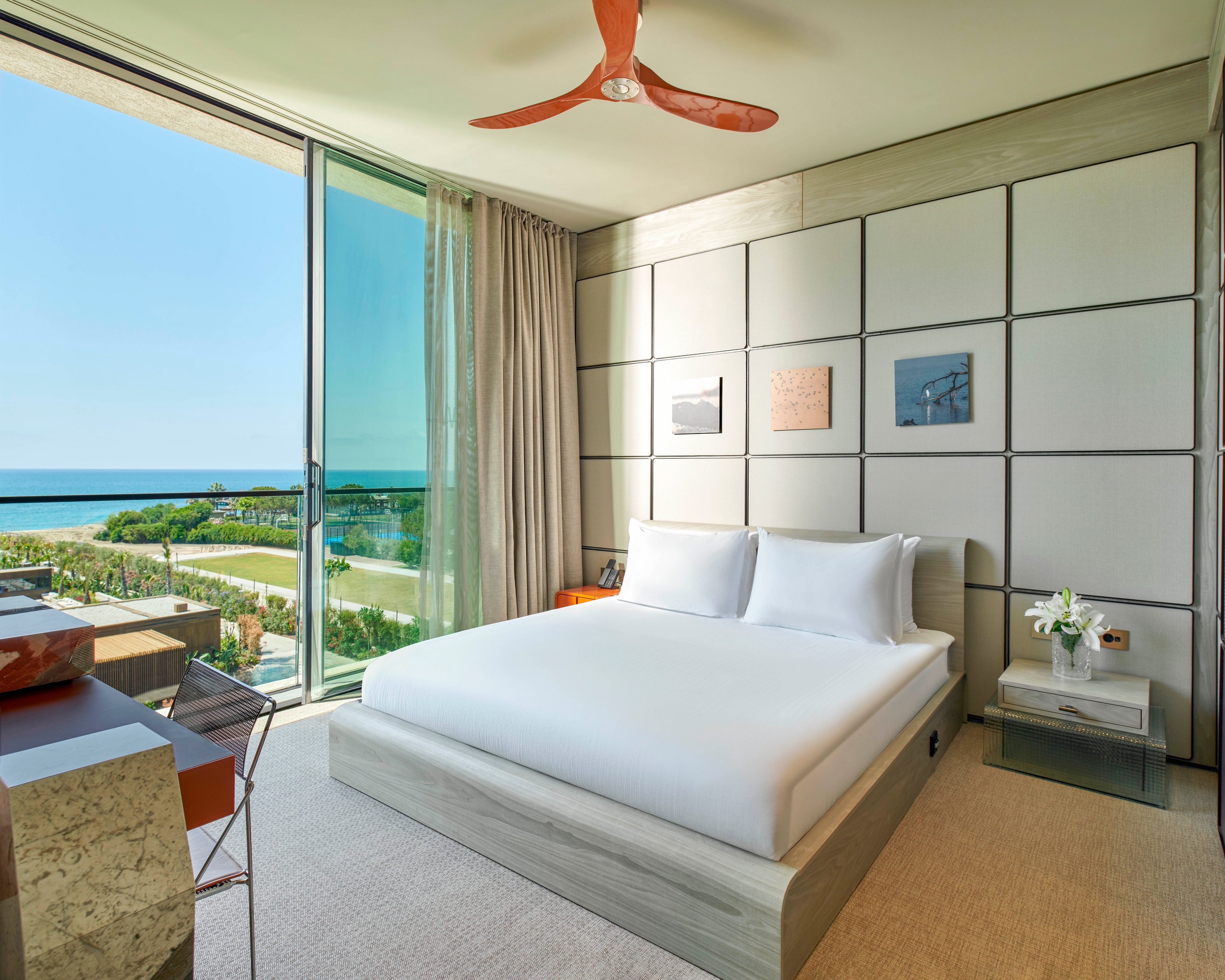 Contemporary hotel bedroom features a large bed with crisp white linens and minimalist furniture. Floor-to-ceiling glass doors open to a balcony overlooking a landscaped area and the ocean. Neutral tones dominate the room, accented by a red ceiling fan and small framed wall art. Soft natural light fills the space, creating a calm and relaxing atmosphere.