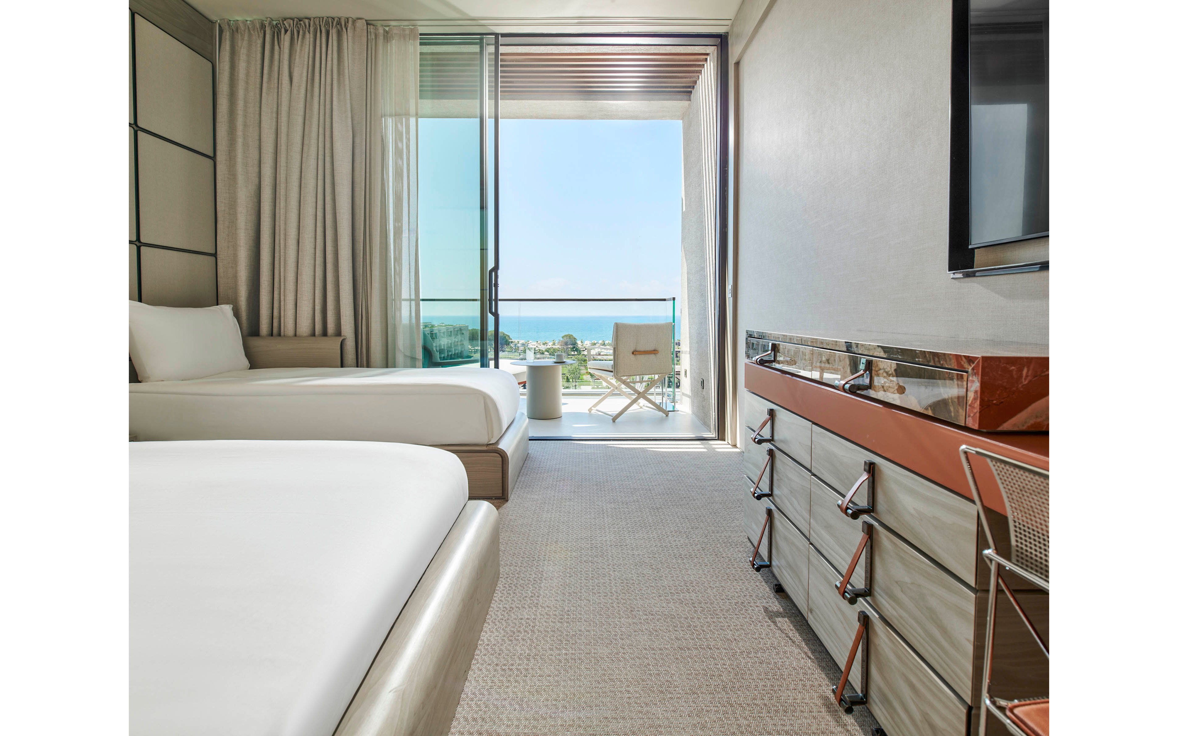 A contemporary hotel room features two neatly made beds and a sleek dresser with a mounted flat-screen TV. Floor-to-ceiling glass doors open onto a balcony with a chair and small table overlooking the ocean and distant coastline. Neutral tones, soft carpeting, and light curtains create a calm, minimalist atmosphere. The space feels bright and airy, emphasizing relaxation and scenic views.