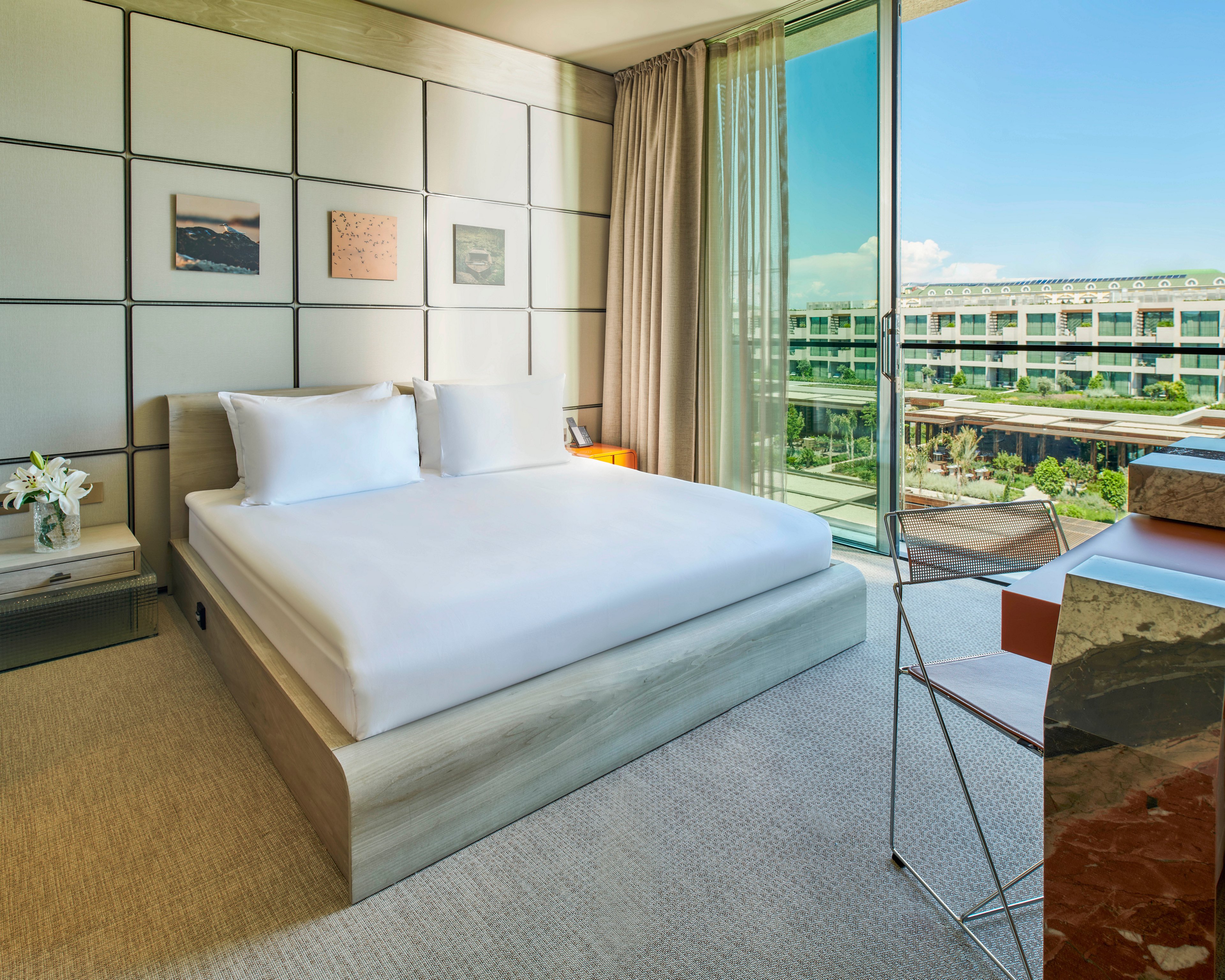 A contemporary hotel bedroom features a minimalist bed with crisp white linens and pillows. Floor-to-ceiling glass windows reveal a view of a modern building and landscaped outdoor area. Neutral tones, soft carpeting, and simple wall art create a calm, upscale atmosphere. A sleek desk and transparent chair add to the clean, modern design of the room.