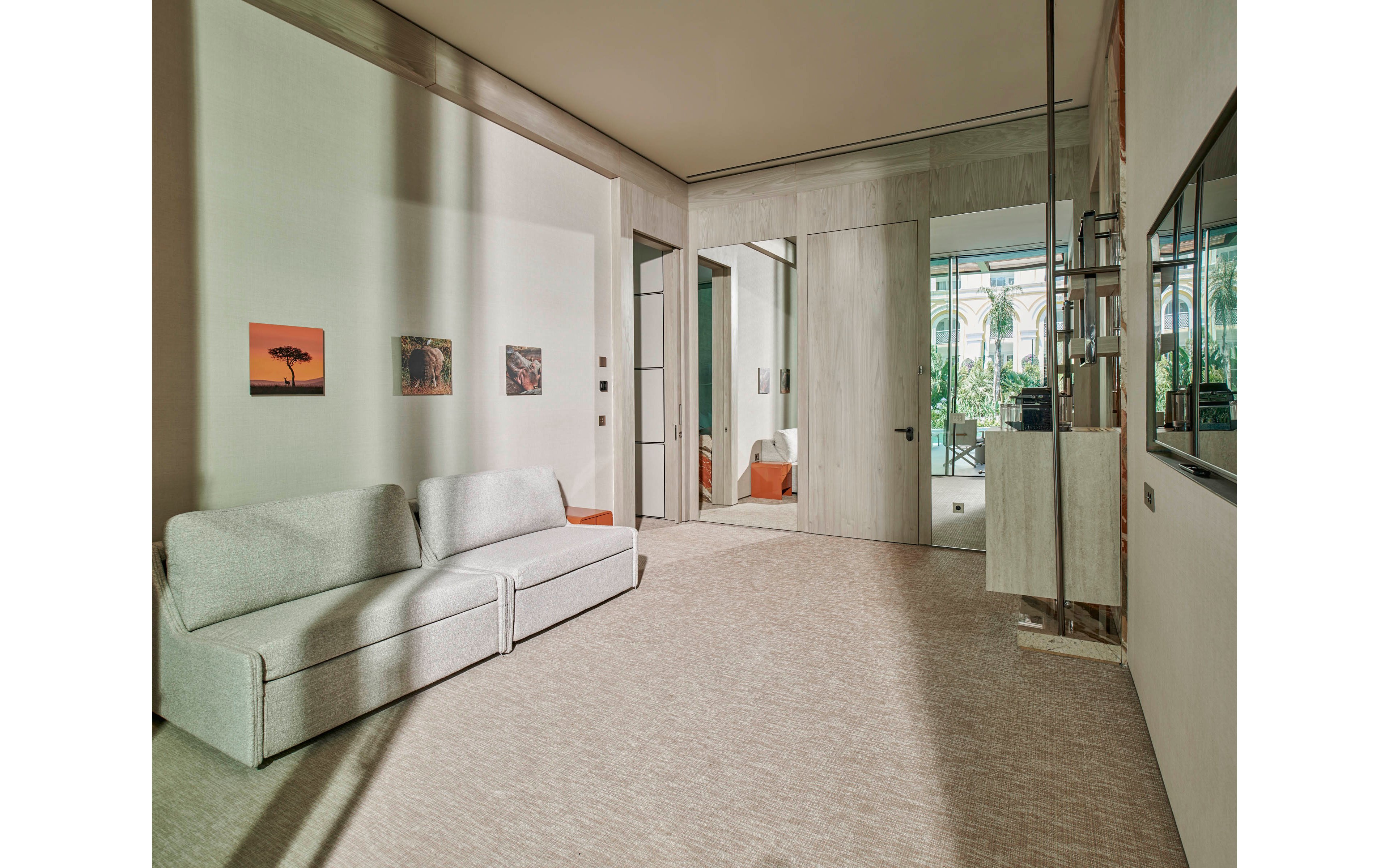 A contemporary living room features a light gray sofa against a neutral wall. The space is decorated with small framed artworks and large floor-to-ceiling doors leading to a bright area with greenery outside. Soft, natural light fills the room, highlighting the clean lines and muted color palette. The overall atmosphere is calm, spacious, and minimalist.