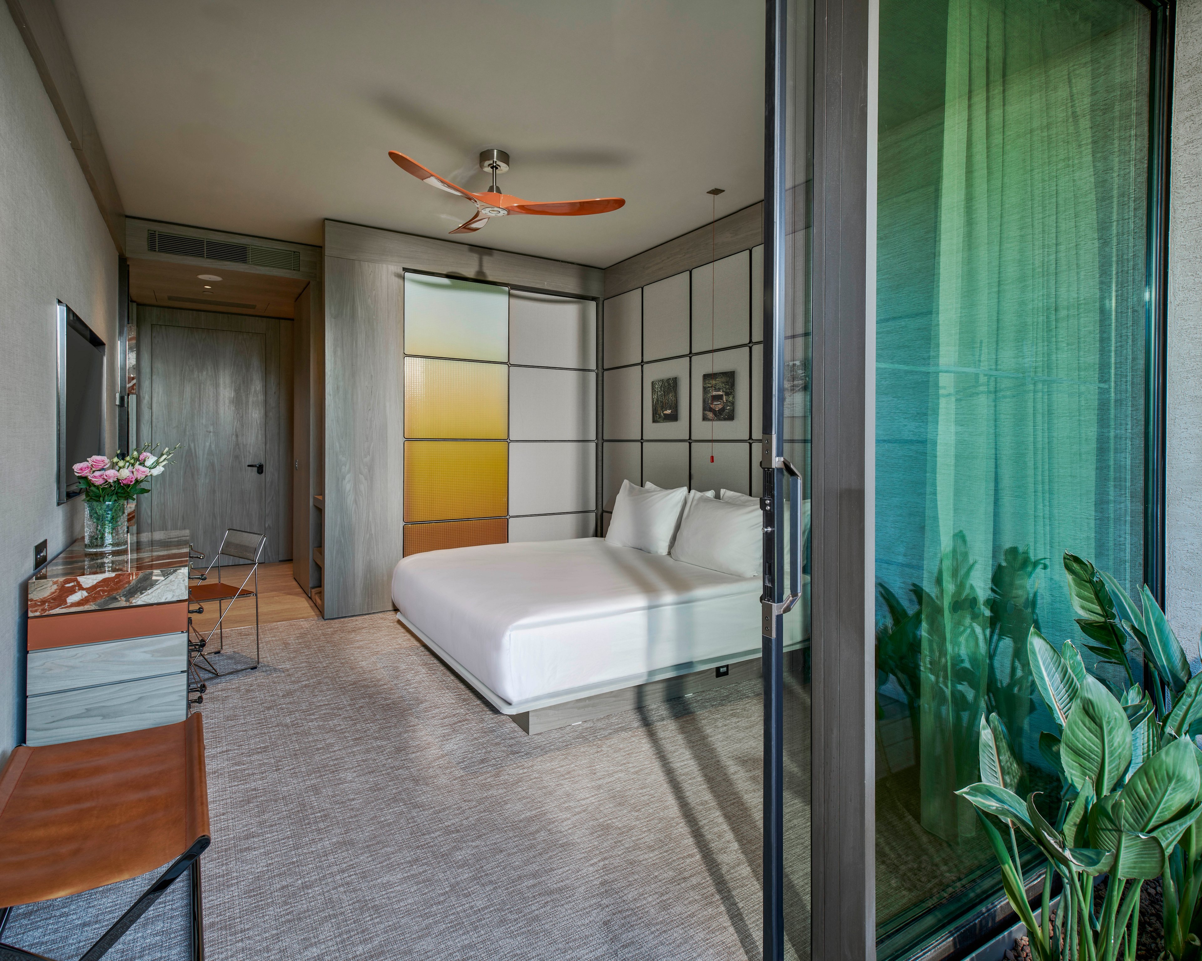 A contemporary hotel room features a low white bed centered against a paneled wall with framed artwork. Sliding glass doors open to a balcony area with lush green plants and sheer curtains. A small desk with flowers, a chair, and a ceiling fan add functional comfort to the minimalist design. Soft natural light and muted tones create a calm, inviting atmosphere.